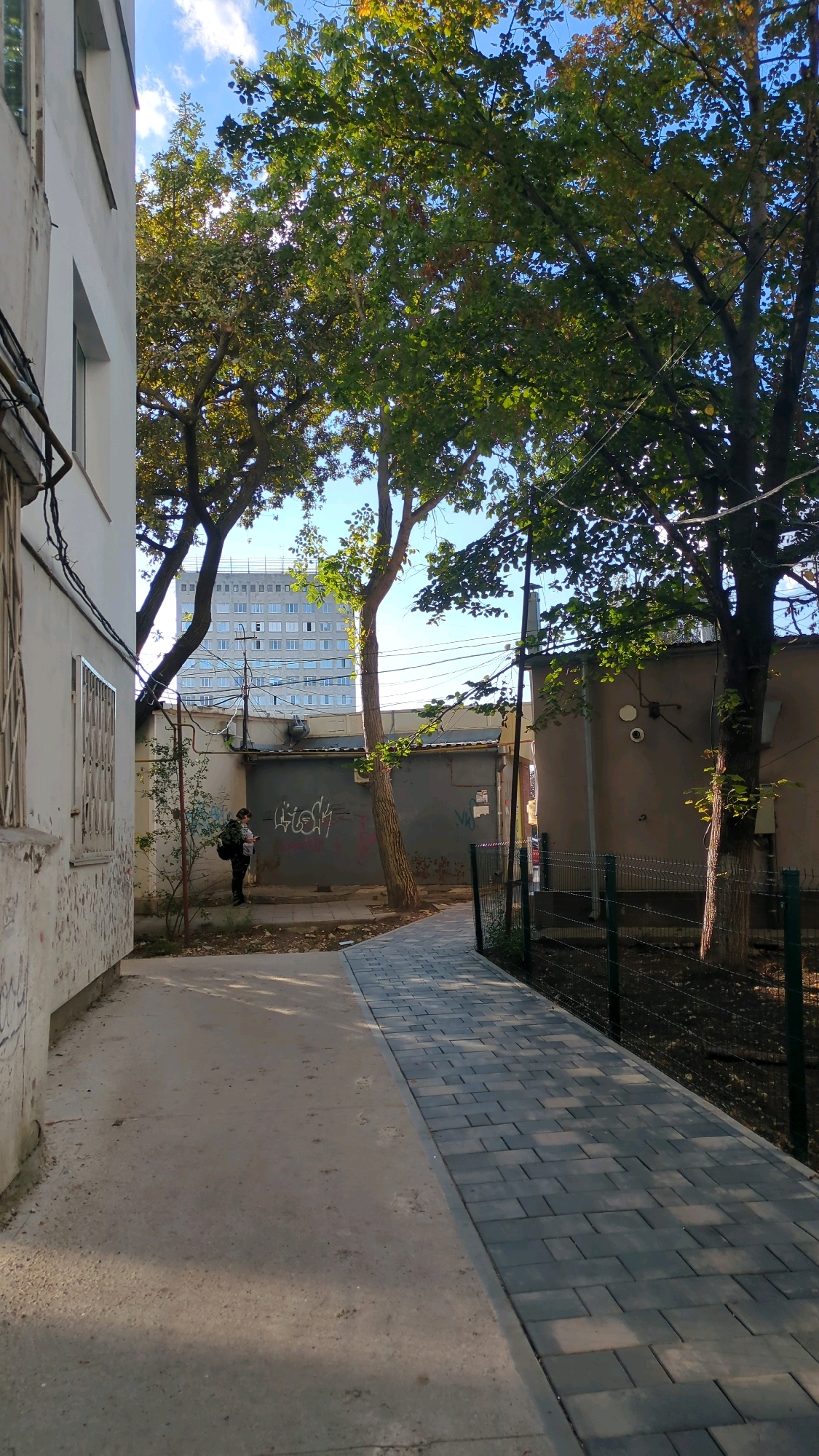 A person wearing a dark backpack and a lighter colored shirt stands near a gray metal garage door with graffiti. The person is looking at a mobile device. The garage is located in an alleyway with a concrete floor and a paved walkway. Trees with green and yellow leaves are present, with a large building in the background. The sky is blue with some clouds. The scene is likely in Chișinău, Moldova, given the architectural style of the buildings and the presence of trees.