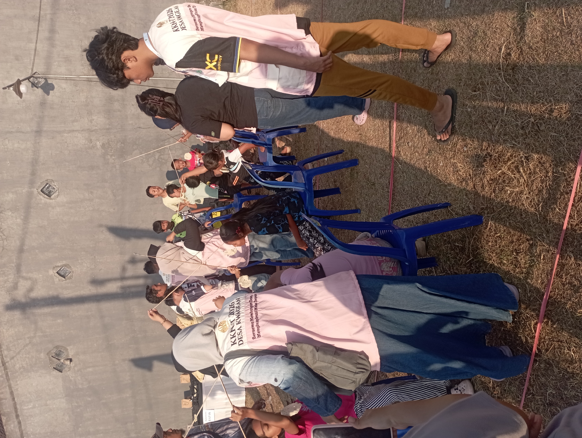 The image depicts an outdoor gathering of people, likely a community event, in Candirejo, Indonesia. The scene takes place in front of a textured concrete wall. In the foreground, a young person wearing a pink vest with the text "KKN T 2026 DESA KWAGEAN" and "Bersama Membangun Desa, Menginspirasi Masa Depan" is visible. This individual is holding a long stick. To the right, another young person in a pink and white shirt with the text "XC NOLOGY" is also present. Several children are actively engaged, holding sticks and interacting with each other. Blue plastic chairs are scattered throughout the area, with some people seated and others standing. The ground is covered with dry grass and straw. In the background, there are tables with items on them, possibly for a market or information booth. Shadows cast by people and objects suggest it is daytime, with sunlight illuminating the scene. The overall atmosphere appears to be one of a community activity or educational program.
