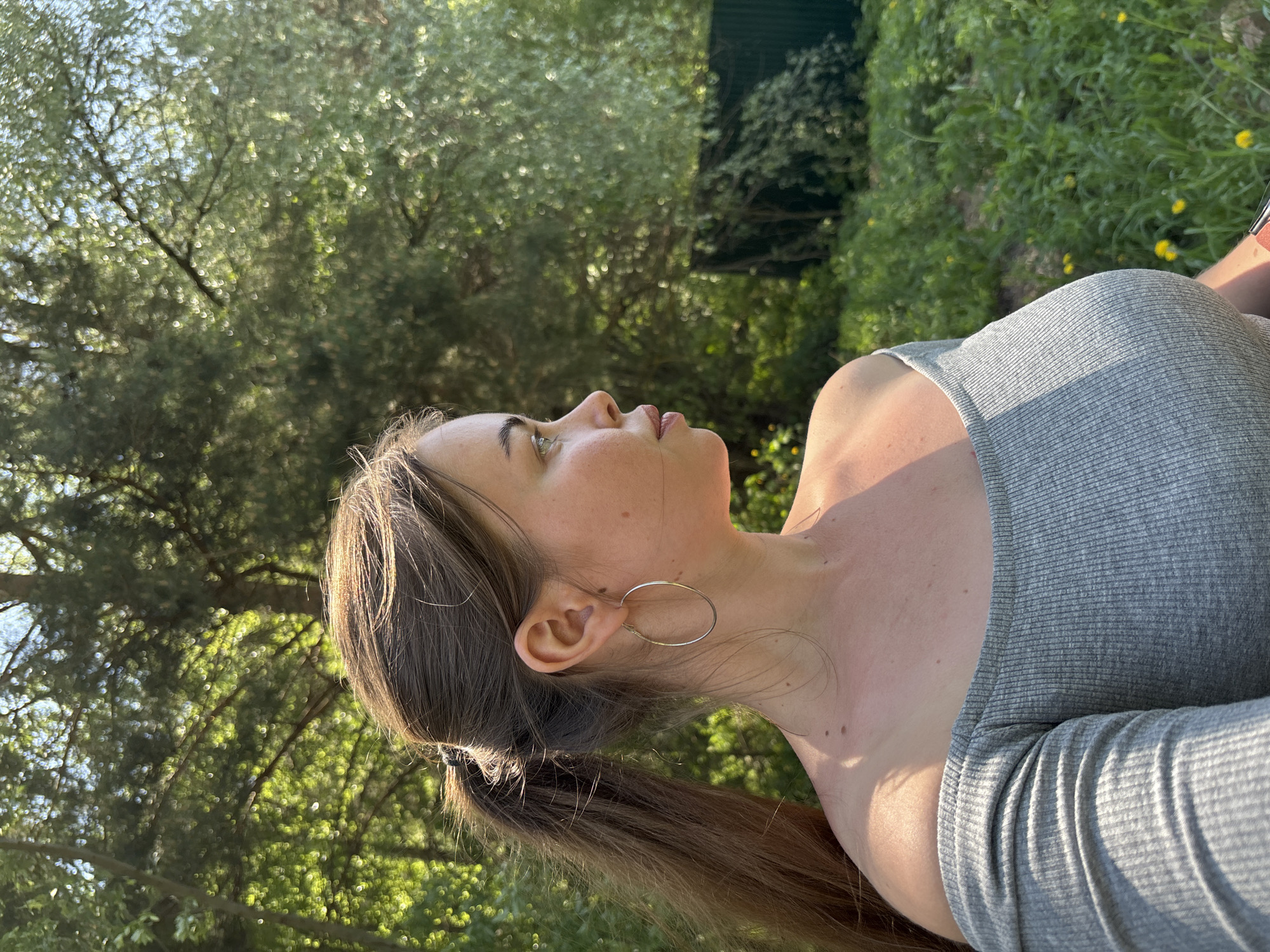 A profile shot shows a woman in a gray shirt with an ear ring on a sunny day. In the background are leafy trees and greenery and a green fence. The location may be in the city of Русский Вожой, Russia.