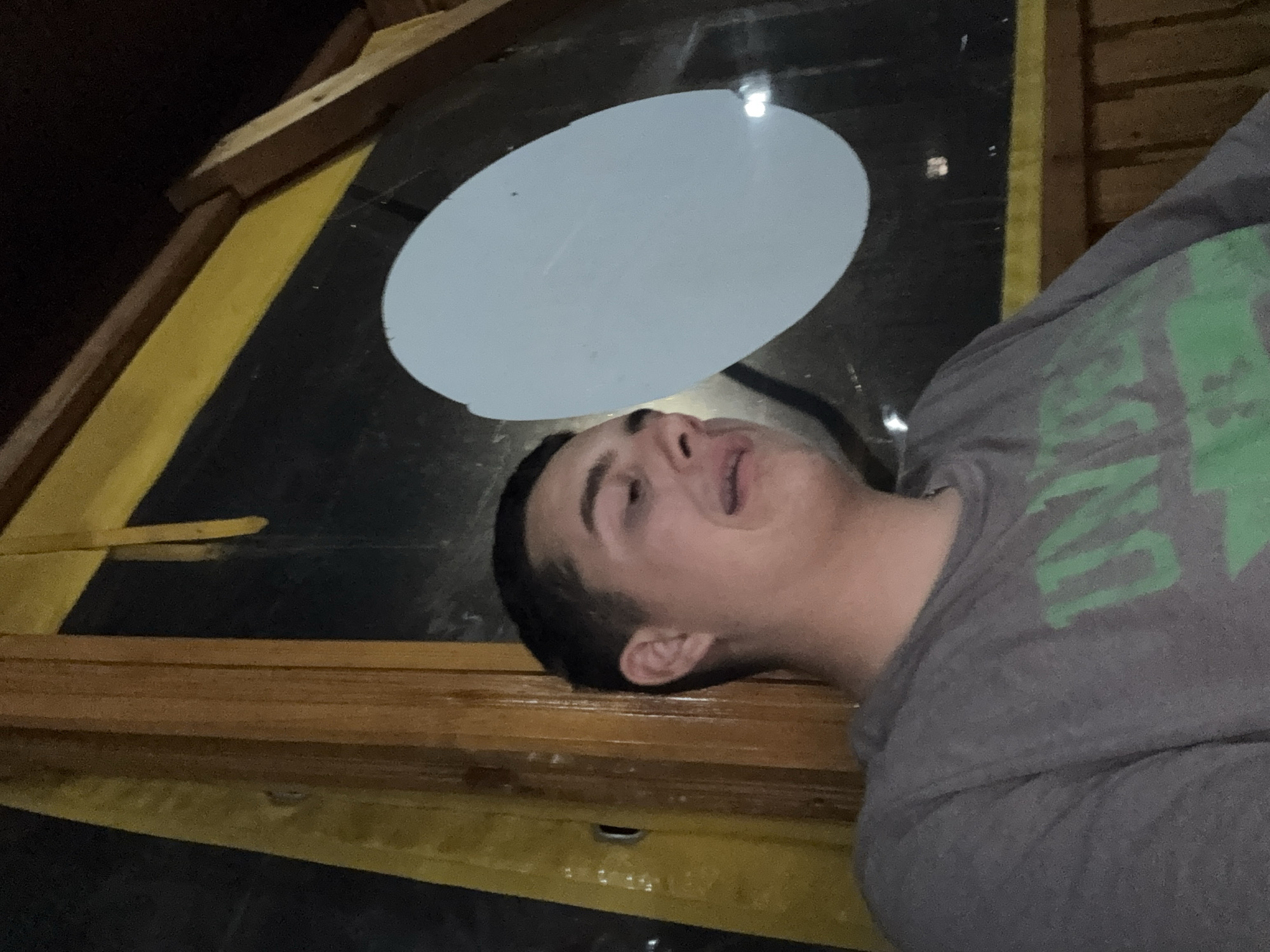 The scene is located in Basarbovo, Bulgaria. A young Caucasian male, appearing to be in his late teens or early twenties, is depicted in a partial profile view from the left side of the frame. He has short, dark hair and is wearing a grey t-shirt with visible green lettering, partially reading "UN". His head is tilted downwards and to the right, with his mouth slightly ajar, suggesting a vocalization or specific facial expression. The subject is positioned within a structure characterized by exposed brown timber framing and yellow paneling/trim, visible vertically on the left and horizontally along the top right. A dark background occupies the upper right and central areas, possibly a window at night or a darkened interior wall. A prominent, opaque white oval object is affixed to or reflecting on this dark background, positioned centrally in the upper right quadrant. Minor glints of light are visible on the dark surface near the oval object.