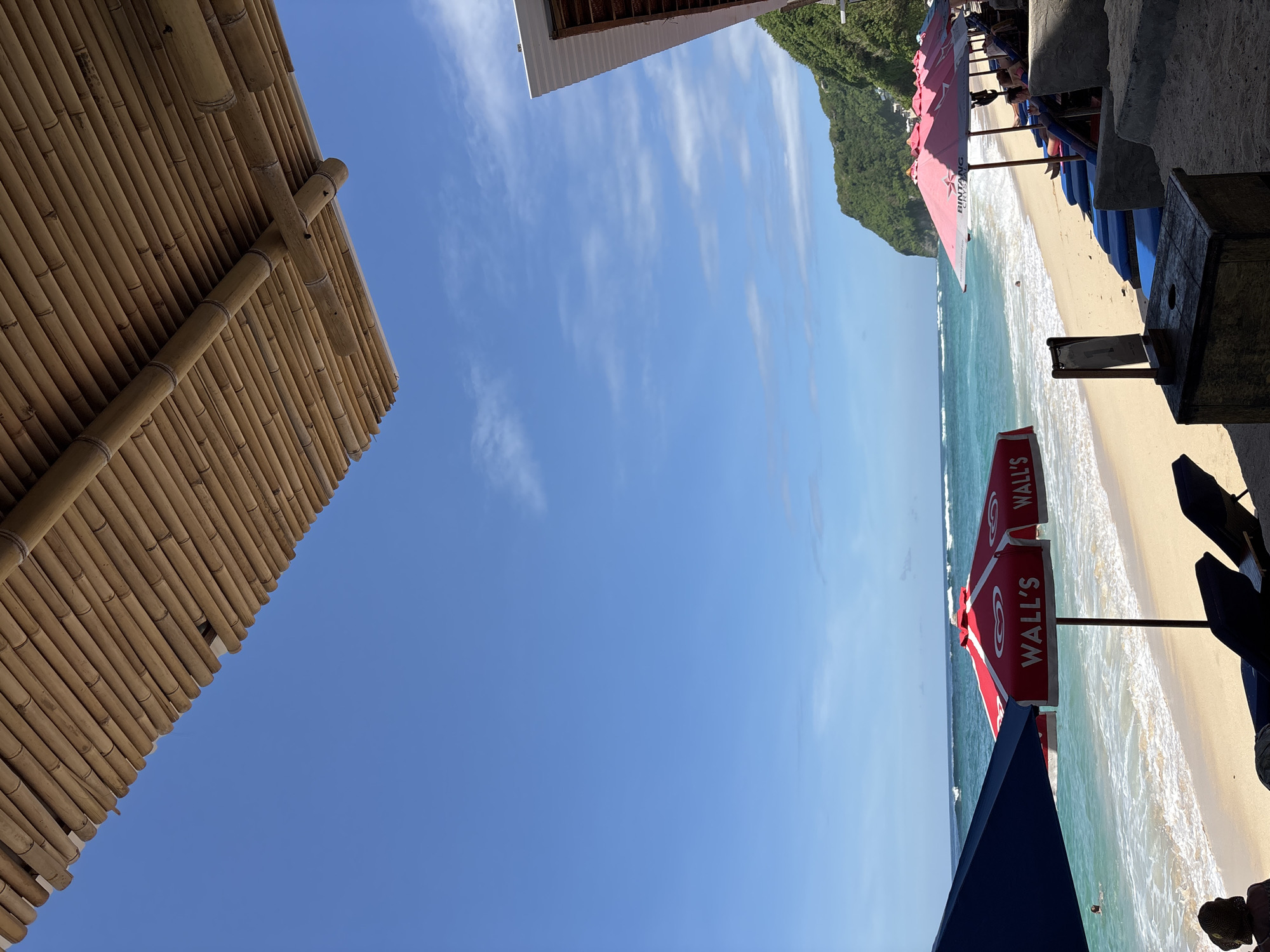 The image shows a beach scene in Kuta Selatan, Indonesia. The view is framed by a bamboo structure on the left and a building's edge on the top. The central focus is the beach with waves, sand, and several beach umbrellas, some red with "Wall's" printed on them and others pink. Some lounge chairs can also be seen on the beach. A green, hilly landscape forms a backdrop to the beach. The sky is blue with wispy clouds.