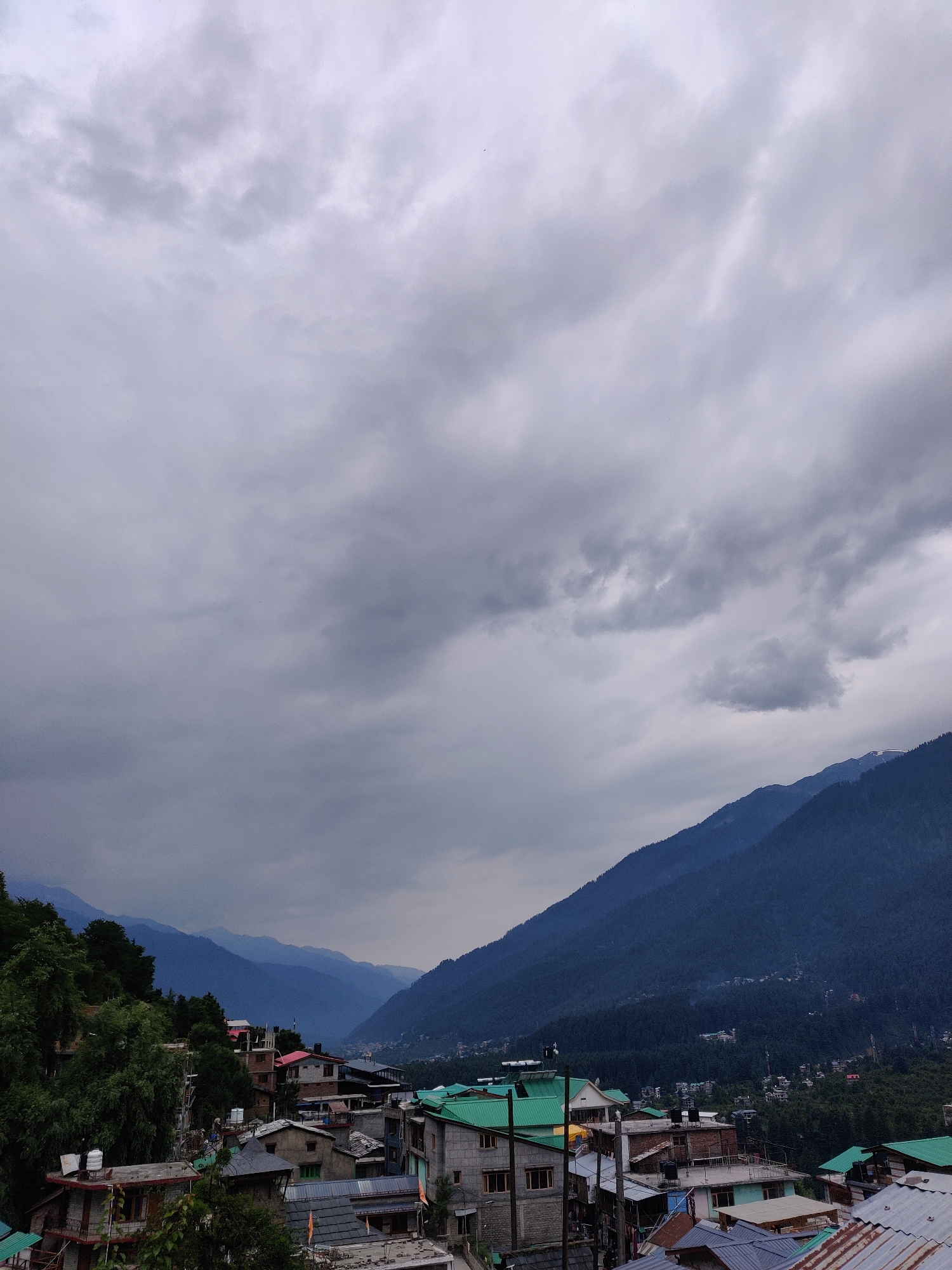 This is a high-angle, full-shot photograph of the town of Vashisht, India, under a cloudy sky. In the foreground, a collection of buildings with various roof types, including green and corrugated metal, are visible. Some buildings have water tanks on their roofs. Lush green trees are interspersed among the structures, particularly on the left side of the frame.
In the midground, the town sprawls into the valley, with more buildings and vegetation. The background is dominated by several layers of mountains, covered in dense, dark green forests. The mountains recede into a hazy blue distance, suggesting a significant elevation and a broad landscape. The sky is overcast with heavy, gray clouds, which cast a muted light over the entire scene. There are no visible people or animals in the image. The overall impression is one of a mountain town nestled within a vast, natural landscape under a dramatic sky.