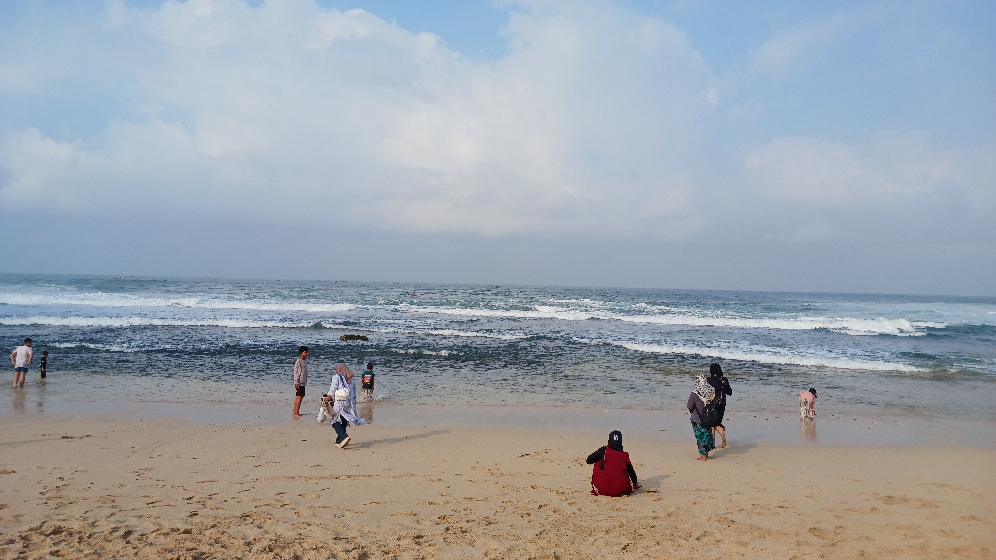 The image depicts a beach scene on a cloudy day in Tanjungsari, Indonesia. The sandy beach is wet and marked with footprints. Several people are present. On the left, two young boys are wading in the shallow water. Further right, a person in a light-colored shirt and dark pants is standing near the water's edge. Closer to the foreground, a woman wearing a white and blue striped top and dark pants, with a white bag over her shoulder, is walking across the sand. To her right, a group of three individuals, two adults and a child, are standing in the shallow water. In the mid-ground of the beach, a person in a red dress is sitting down, facing away from the camera. In the distance, two small boats are visible on the ocean. The ocean has moderate waves breaking near the shore, and the sky is a mix of blue and white clouds.
