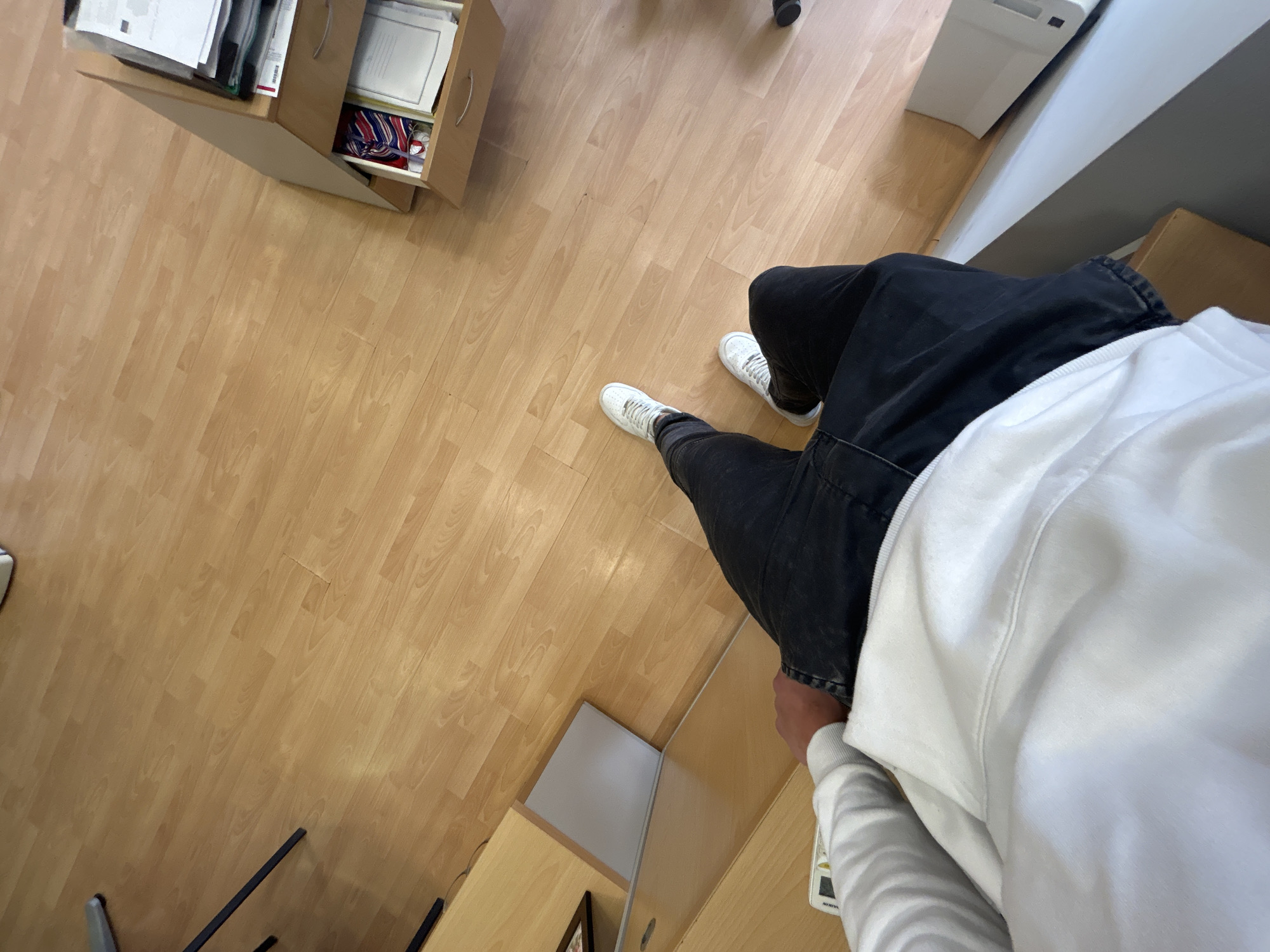 A first-person perspective photograph shows a person wearing black jeans and white sneakers standing on a light-colored wooden floor. The person is also wearing a white sweatshirt. A filing cabinet is visible in the top left corner of the frame. A desk and a white printer are located on the right side of the person. The location appears to be an office setting. The city of Zlatograd, Bulgaria, cannot be determined from the visual information provided.