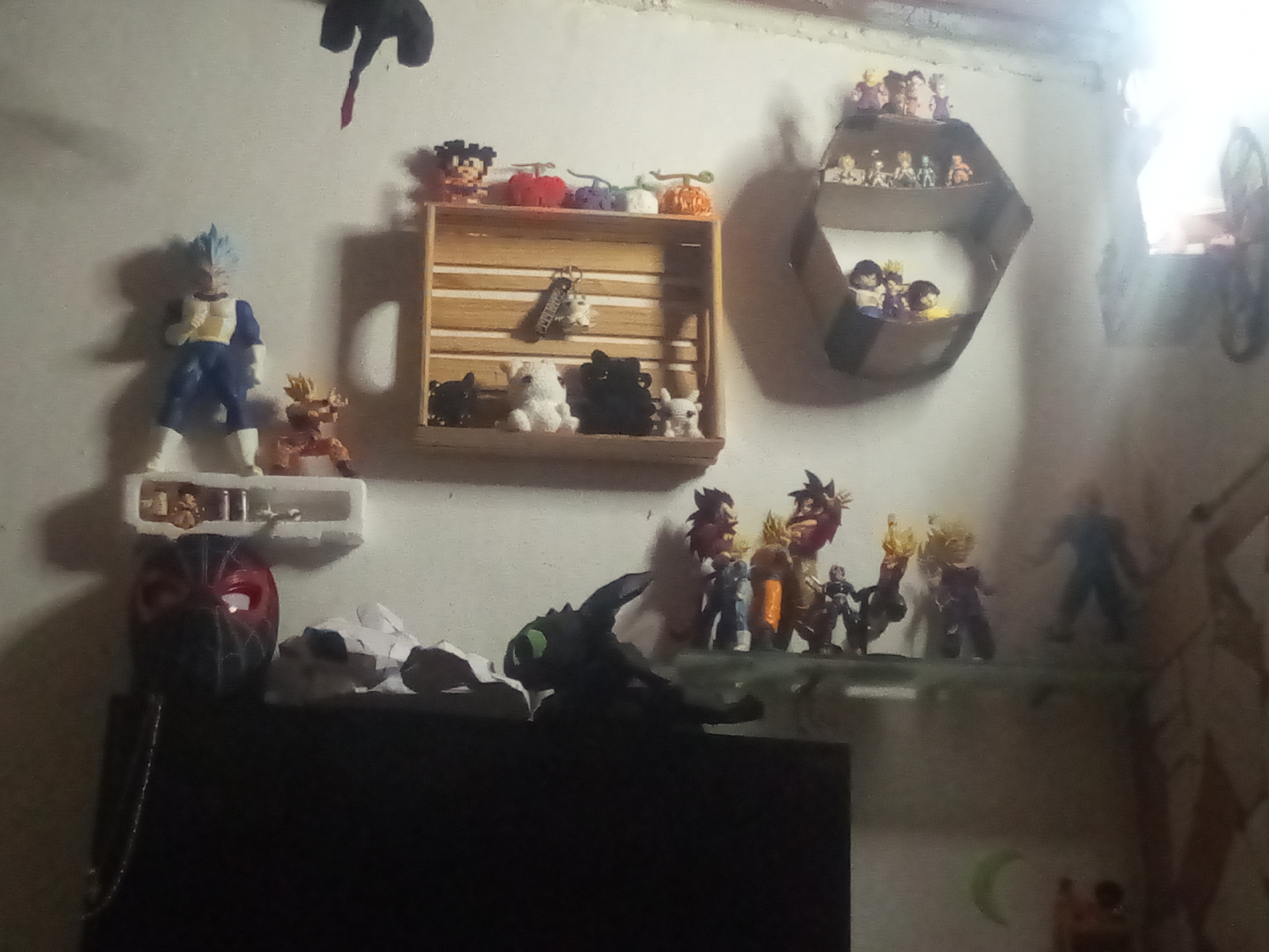 The image depicts a room interior, cluttered with various figurines and decorative items, likely a personal living space. The location is El Valle del Espíritu Santo, Venezuela. The walls are off-white and serve as backdrops for various displays.
On the left, a shelf holds a blue-haired figure in a battle stance, likely a character from Dragon Ball, accompanied by a smaller figure. Below the shelf, a red mask, perhaps related to the character Deadpool, sits atop a dark surface, possibly a TV or cabinet. To the right, a wooden crate is mounted on the wall, containing small animal figurines and knitted items resembling fruits. A hexagonal shelf above it contains more figurines.
Across the middle of the image, a line of Dragon Ball figurines stands on a shelf, including a dark dragon-like figure with glowing green eyes in the foreground. To the right, a blue figure stands alone on another shelf.
The image appears to be lit by natural light coming from a window on the right, which partially obscures a bicycle hanging on the wall. The overall environment is cluttered and personal.