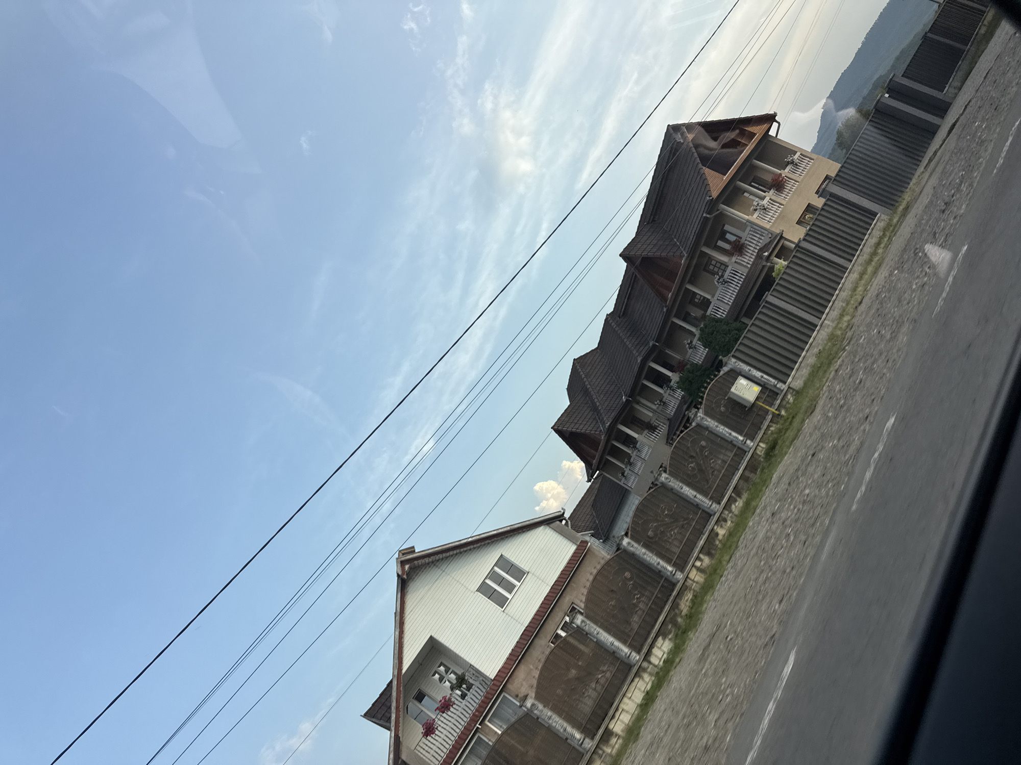 In Oituz, Romania, two residential houses are depicted from a low-angle, roadside perspective. The house on the left is white with brown trim and a balcony on the upper floor. It has a peaked roof and a large fenced yard. The house on the right is a tan color with a darker roof and multiple balconies. It is also fenced. A road runs in the foreground, and power lines crisscross the sky. In the background, hills and mountains are visible, with a cloudy sky above. The scene appears to be set during daylight hours.