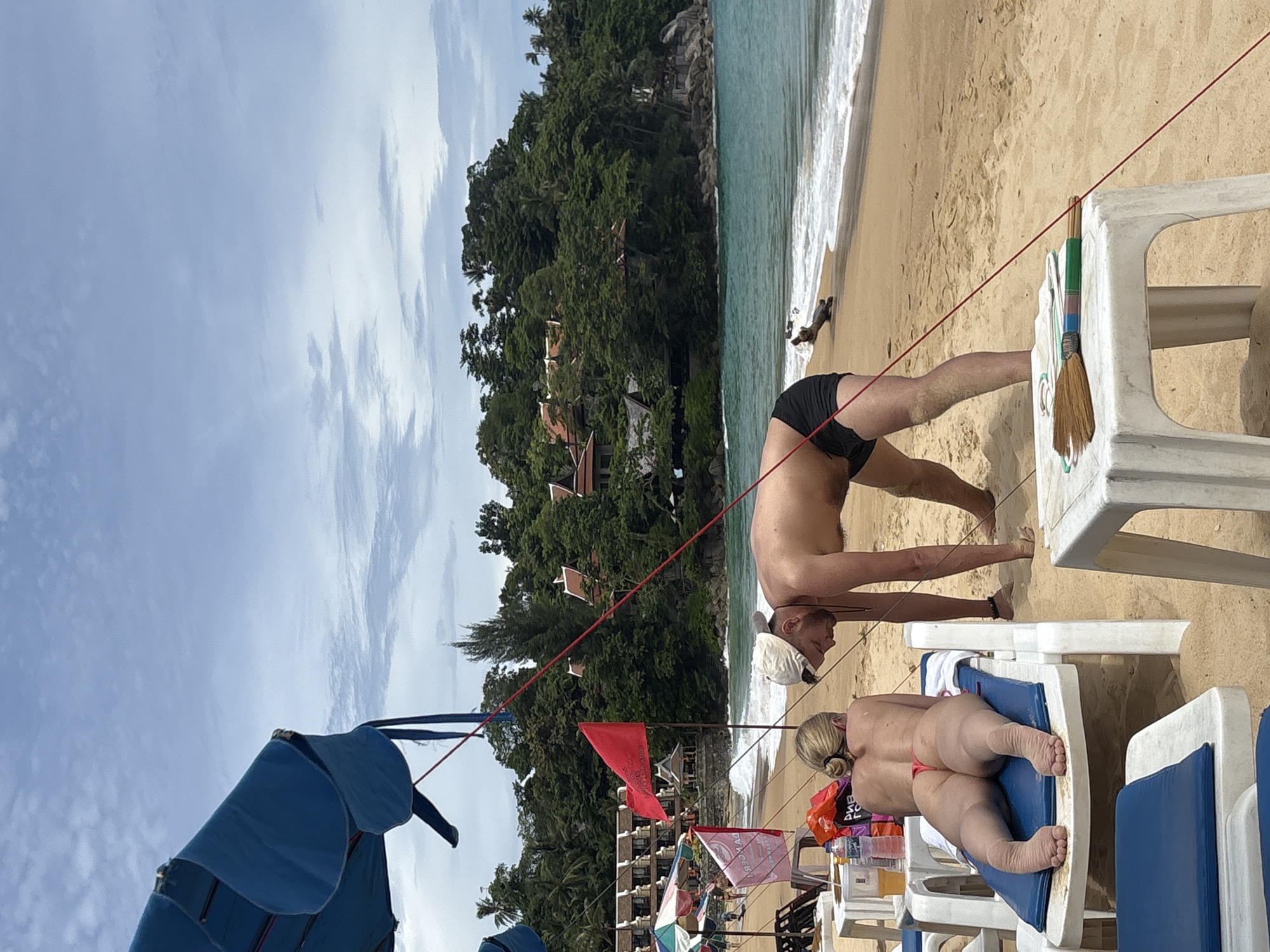The image depicts a beach scene in Karon, Thailand. In the foreground, a woman lies on a blue lounge chair, facing away from the camera, wearing a red thong. A man, wearing black swim trunks and a white cap, is standing next to her, bent over. In the middle ground, a red flag flies from a pole, and a pink flag is visible behind it. Further back, there are several beach umbrellas and buildings nestled amongst lush green trees on a hillside overlooking the turquoise ocean. The beach is sandy, with waves gently rolling onto the shore. A small piece of driftwood is visible on the wet sand near the water's edge. The sky is partly cloudy. A small brush and some cleaning supplies are on a white plastic table in the foreground.