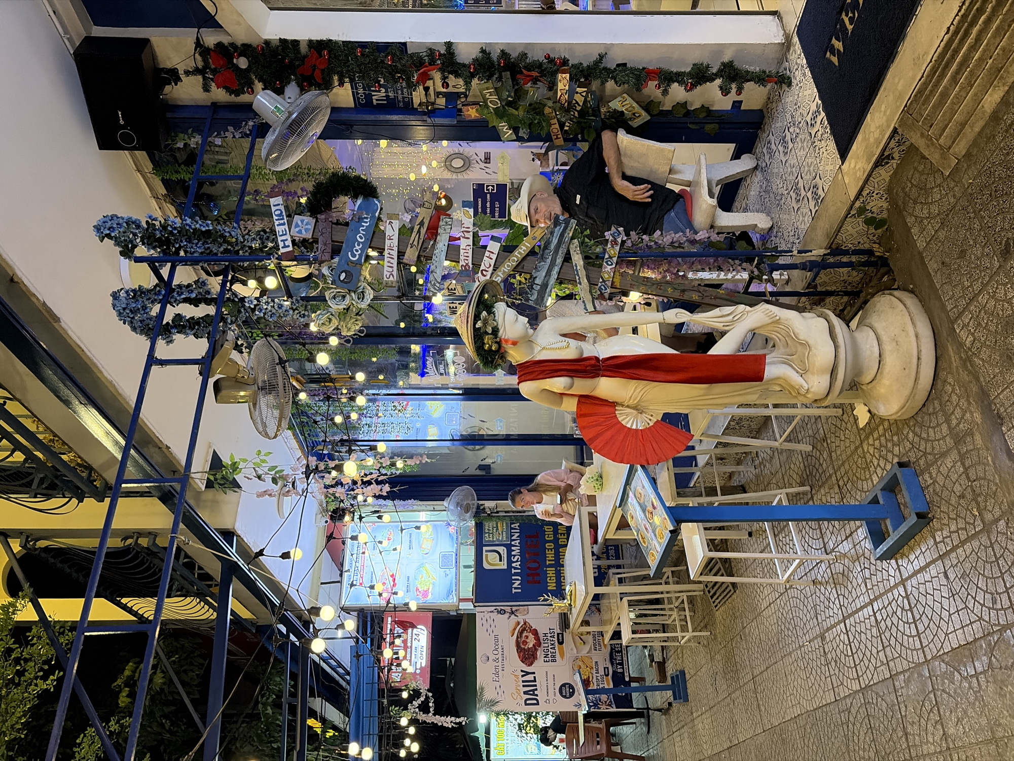 The image depicts the exterior seating area of the Eden & Ocean Restaurant in Đà Nẵng, Vietnam, which also serves as the TNJ Tasmania Hotel. The scene is illuminated by strings of hanging lightbulbs and decorative lights, creating a festive ambiance. In the foreground, a white statue of a woman with a red fan stands next to a table displaying a menu. A man wearing a cowboy hat and a black shirt is seated at a table to the right, looking towards the camera. Behind him, a woman in a pink top is seated at another table, holding a drink. The restaurant features blue and white accents, with various decorative signs and plants adorning the facade. The tiled flooring and overhead structures suggest an urban street-level establishment.