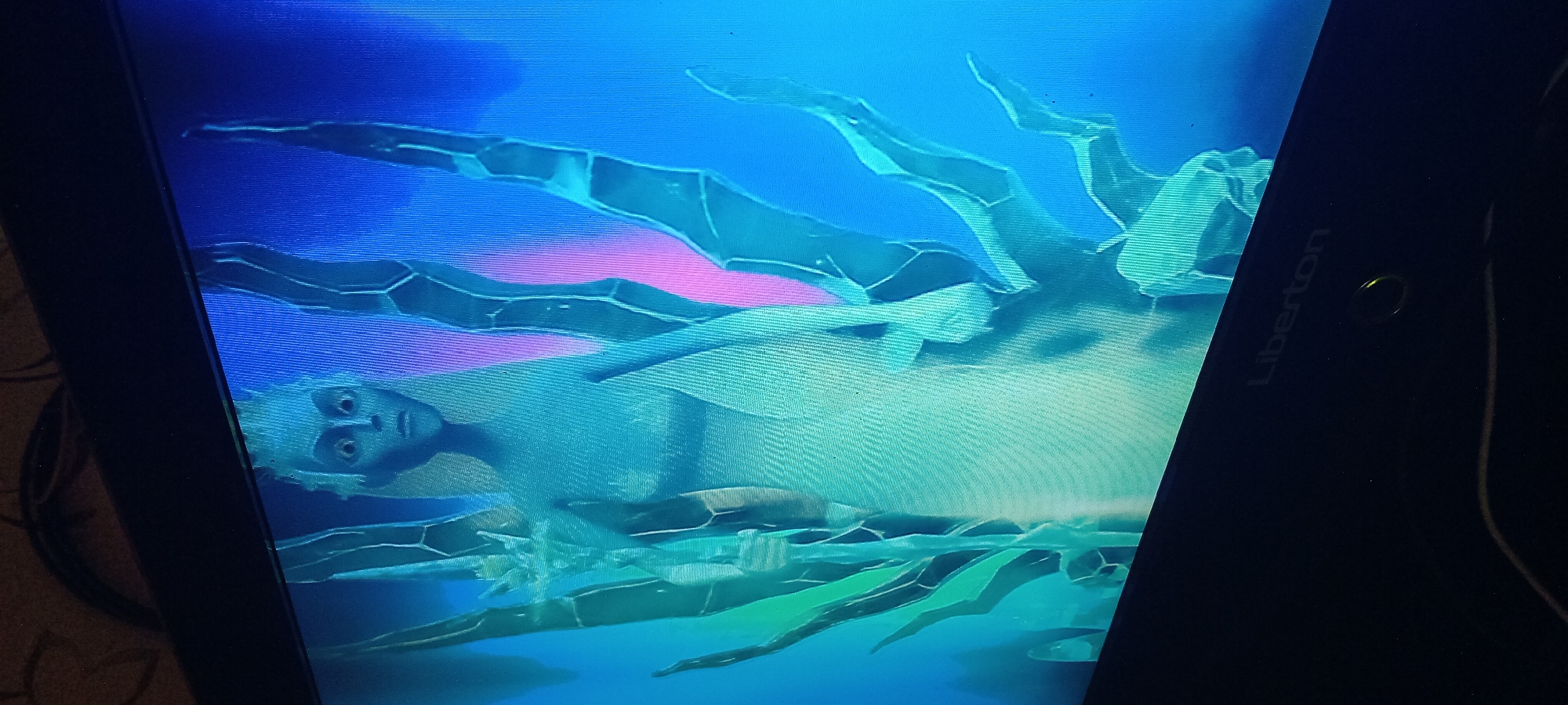 The image displays a still frame from a video game or animation, featuring a pale-skinned, humanoid character with wide, startled eyes and spiky, white hair, positioned centrally. The character appears to be a female figure, wearing a light-colored, flowing garment, and holding a staff-like object in her left hand. She is surrounded by abstract, crystalline or ice-like structures in shades of green and blue, which branch out around her. The background is a vibrant gradient of blue and pink. The screen itself is visible, with a black bezel and the "Liberton" logo subtly printed on the lower right corner. The image is captured from a slight angle, suggesting it is a photograph of a monitor or television screen. There is no indication of the city of Koviaghy, Ukraine, or any urban environment.