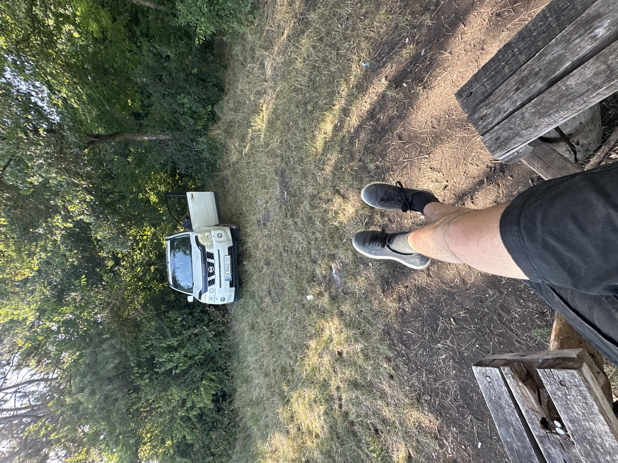 In the city of Velykozalissia, Ukraine, a white Nissan X-Trail SUV with license plate BX 8662 IC is parked on a dirt and grass clearing surrounded by dense green foliage and trees. The driver's side door of the vehicle is open. In the foreground, a person's legs, clad in black shorts and dark sneakers with gray socks, are visible, with their feet resting on two wooden benches. The lighting suggests it is daytime, with sunlight filtering through the trees and casting shadows on the ground.