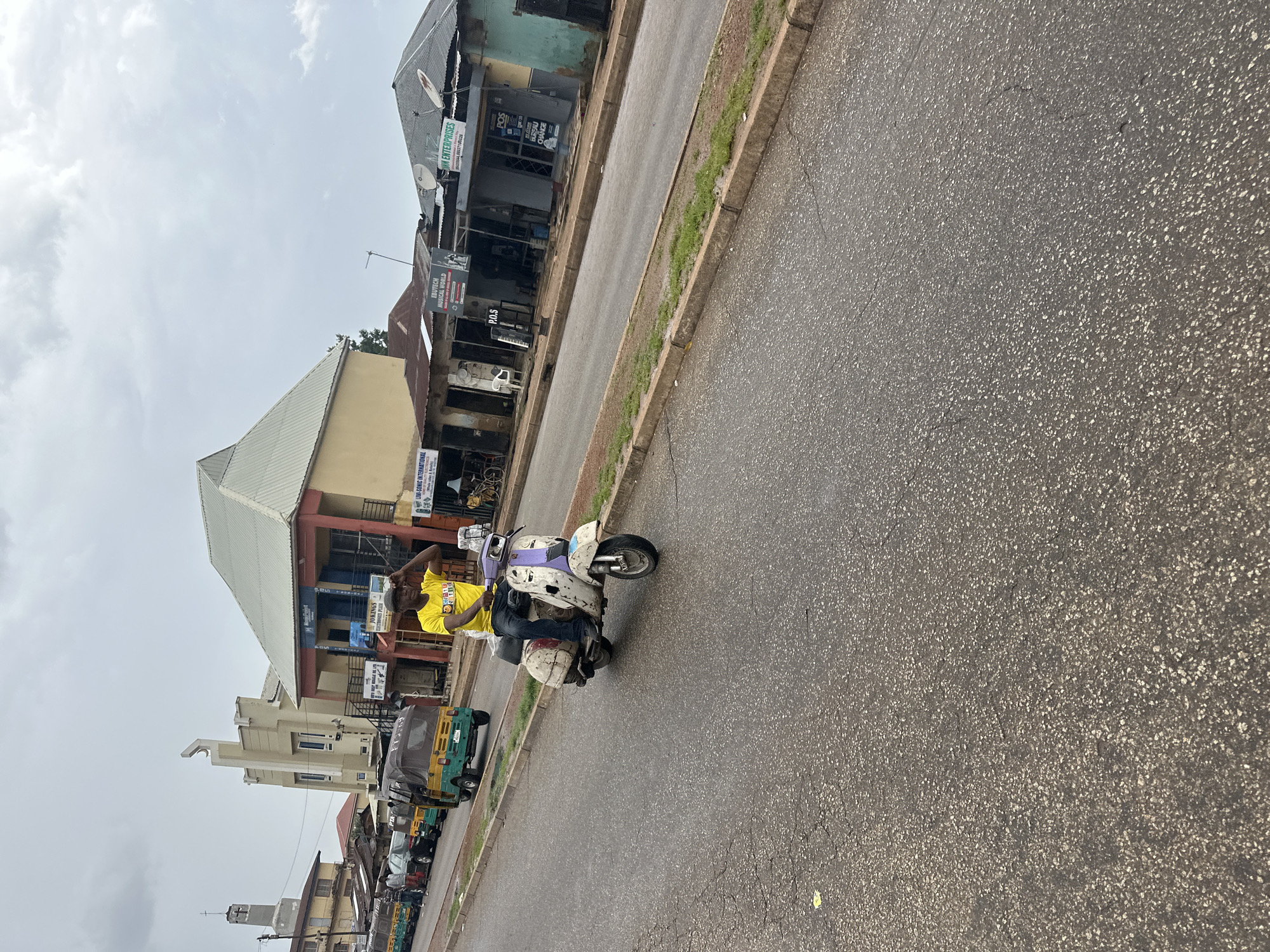A person rides a scooter down a paved street lined with buildings. The scooter, a light purple and white Vespa, has a person in a bright yellow t-shirt and blue jeans riding it. The person appears to be looking to their right, with their left hand touching their head. The scooter has a white box attached to the back. Behind the scooter, several auto-rickshaws, also known as "keke napep," are parked or moving along the street. The buildings on the left side of the street are a mix of commercial establishments, with signs for "JOKINGS," "LIVI-SONIC INTERNATIONAL," "EBUTECH MUSICAL WORLD," "M ENTERPRISES," and "P.O.S." The sky is overcast, suggesting a cloudy day. The scene appears to be in a busy urban area, possibly a market or commercial district in Jos, Nigeria. The time of day is unclear due to the overcast sky, but it seems to be daytime. The overall atmosphere is that of a typical day in a bustling African city.