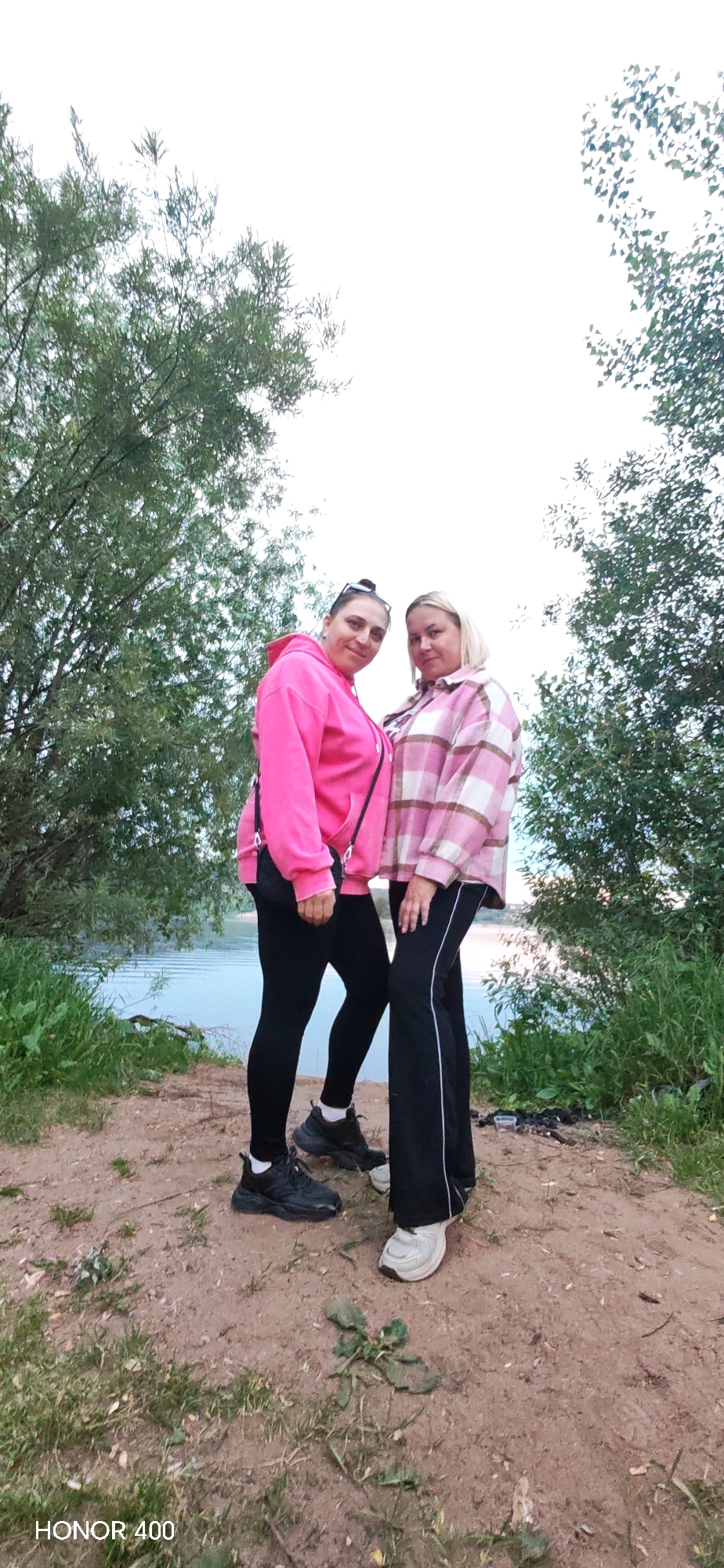 The image shows two women standing on a dirt path near a body of water. The woman on the left is wearing a bright pink hooded sweatshirt and black leggings, with black sneakers. She has a small black crossbody bag. The woman on the right is wearing a pink and white plaid shirt, black pants with a white stripe down the side, and white sneakers. They are standing close together, facing the camera. Trees with green foliage are visible behind them, and the sky is overcast. The water appears to be a lake or river. The location is indicated as Noviy, Russia, by the "HONOR 400" watermark, which is a phone model, not a location.