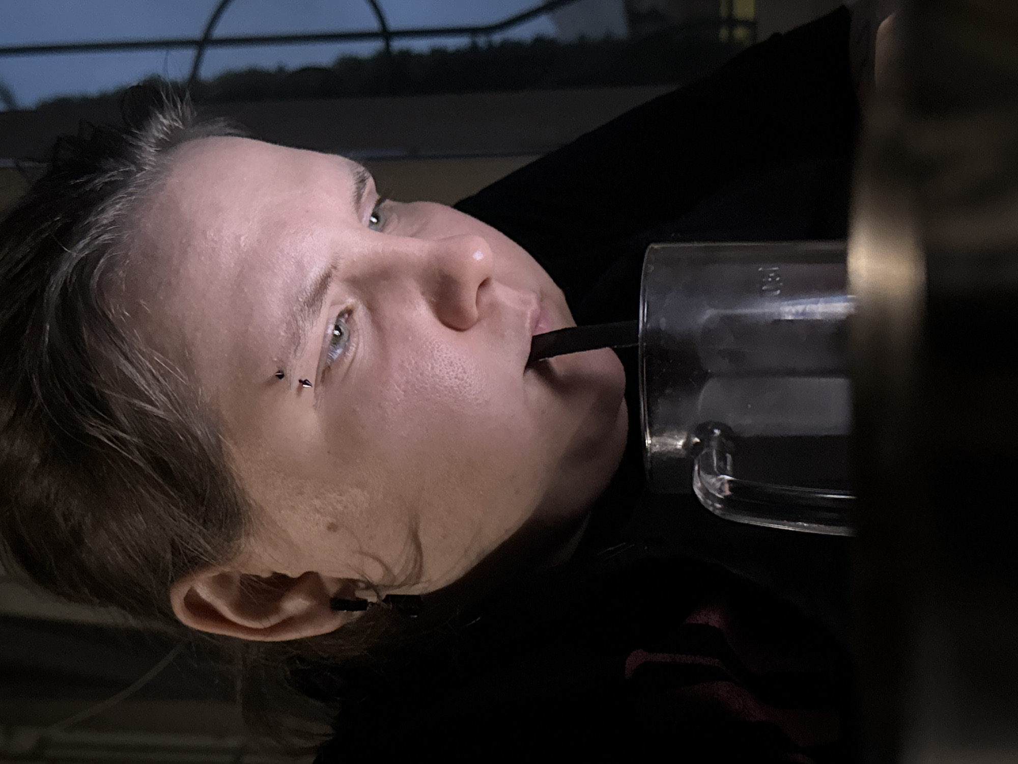 A close-up, waist-up shot shows a person with light skin, light-colored eyes, and dark hair, possibly dyed, with bangs. They are wearing a black shirt with a red logo visible on the left side. The person has a piercing above their left eyebrow. They are holding a clear glass with a black straw and appear to be drinking from it. The background is dark, with some blurry elements suggesting an indoor setting with a window or reflective surface. The lighting is dim, with a light source illuminating the person's face from the side. The overall atmosphere is somewhat moody and introspective. The image is cropped in a way that focuses on the person's face and upper body, with the foreground and background blurred, creating a shallow depth of field. The image appears to have been taken in the city of Zaokskiy, Russia, though there are no specific visual cues in the image itself to confirm this location.