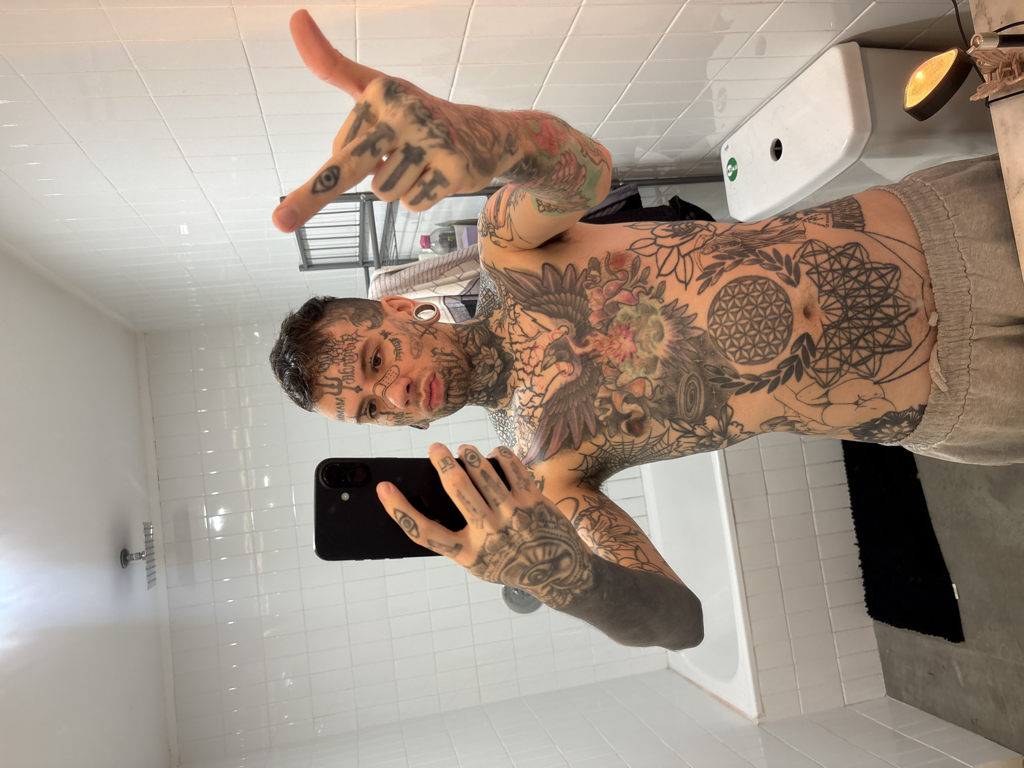 A light-skinned male with extensive tattoos covering his face, torso, arms, and hands is taking a selfie in a bathroom. He is holding a black smartphone in his left hand and gesturing with his right hand. He is wearing grey bottoms and his torso is bare. The bathroom has white tiled walls, a bathtub, a shower head, and a white sanitary unit above the bathtub. A black bath mat is on the floor next to the bathtub. The setting appears to be a standard bathroom, but there are no specific indicators tying it to Buenos Aires, Argentina.