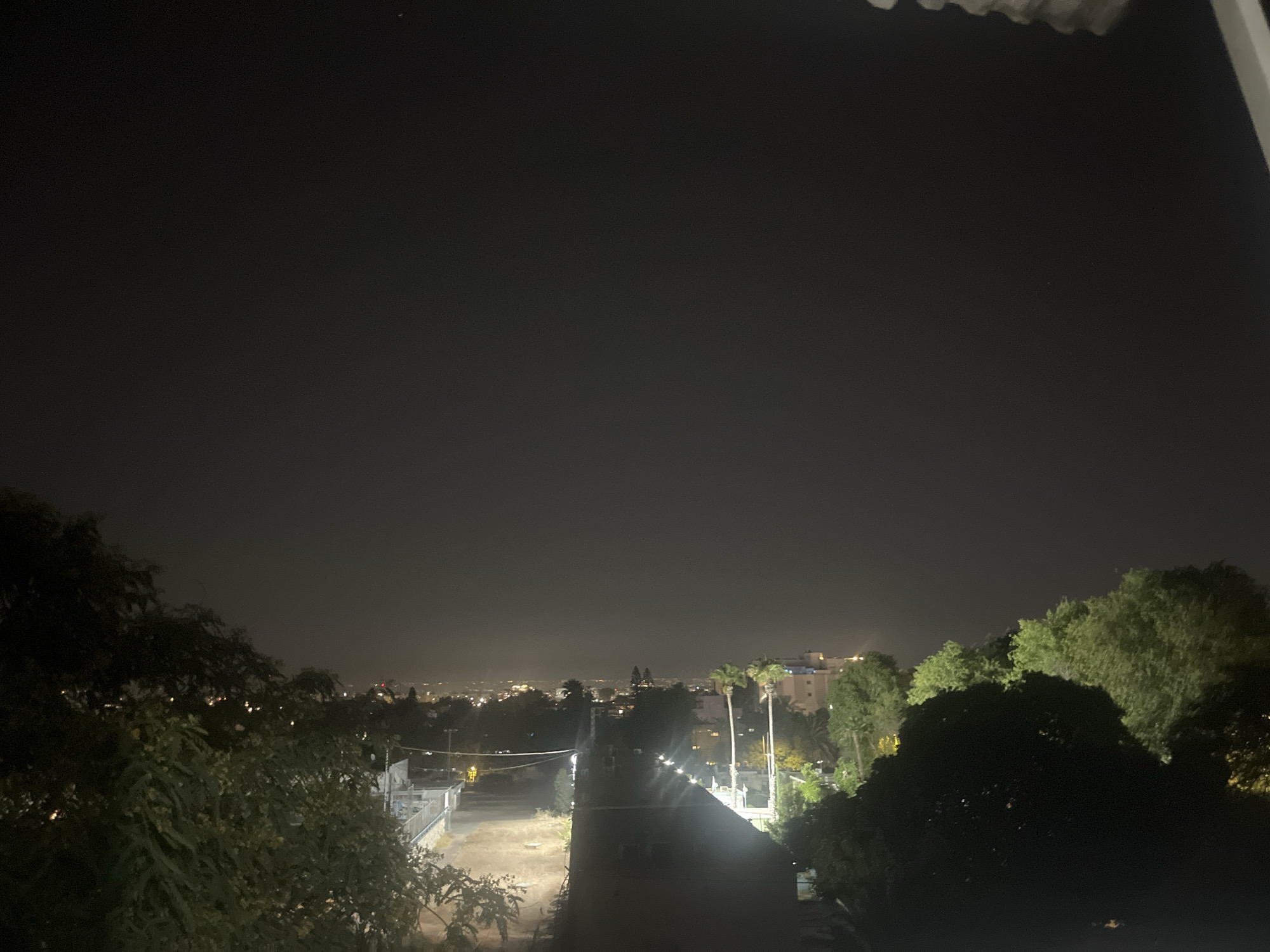 A nighttime aerial view from a vantage point overlooking the city of Ashdod, Israel, showcases a sprawling urban landscape illuminated by artificial lights against a dark sky. The foreground is dominated by lush green trees and foliage, casting deep shadows that obscure details. A paved pathway or road extends into the midground, marked by streetlights casting pools of light. In the distance, the city lights form a dense, glowing tapestry, with scattered buildings and structures visible. The overall impression is one of urban sprawl viewed from above at night, with natural elements framing the man-made environment.