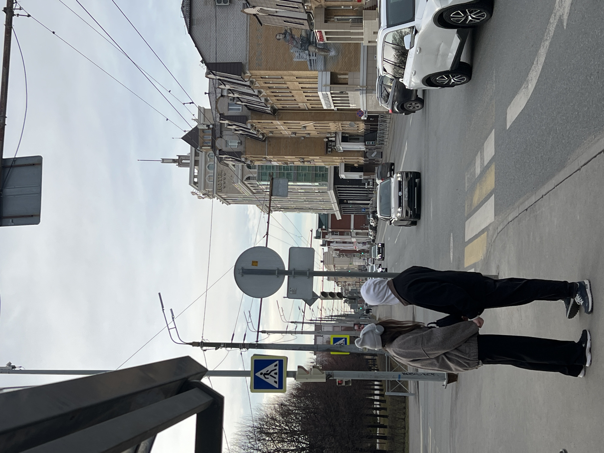 A street scene in Kazan, Russia, features two pedestrians, one in black and one in brown, standing on a sidewalk. In the background, a street with several cars is seen. Street signs and traffic lights are visible along with buildings and overhead wires. The sky is overcast. The building in the background is likely the Shalyapin Palace Hotel.