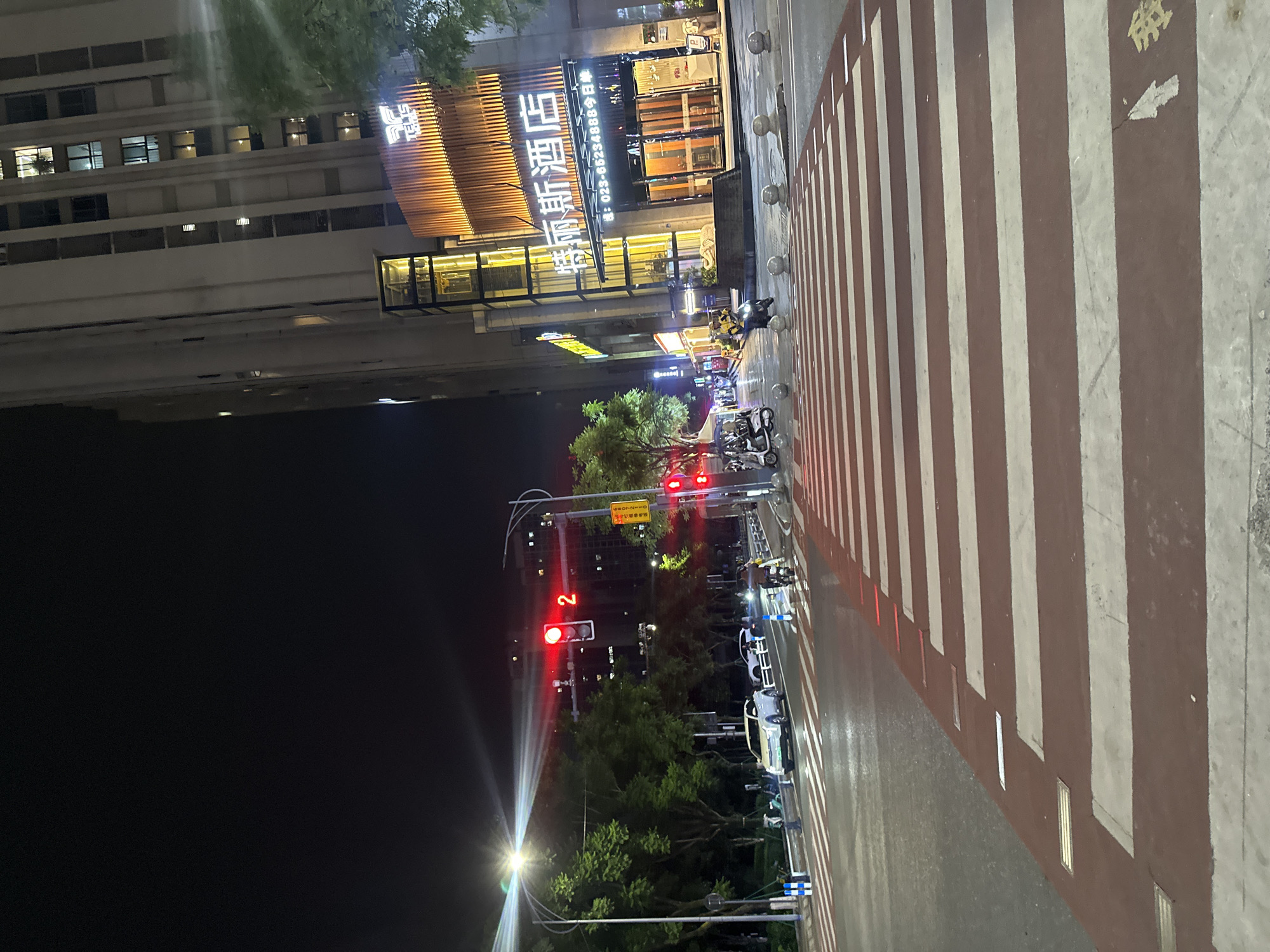 A nighttime scene depicts a street with a crosswalk. The crosswalk, marked with red and white stripes, occupies the foreground, with a portion of a building and a tree visible on the left. The building features illuminated windows and signage, suggesting it's a commercial establishment, possibly a hotel, with Chinese characters and a phone number visible. Several motorcycles are parked along the street, and a few people can be seen near them. Traffic lights are illuminated, with a red light indicating a stop. The overall ambiance is urban and nocturnal, illuminated by streetlights and the building's artificial lighting. The setting appears to be a relatively modern area in Chongqing High-tech Zone, China.