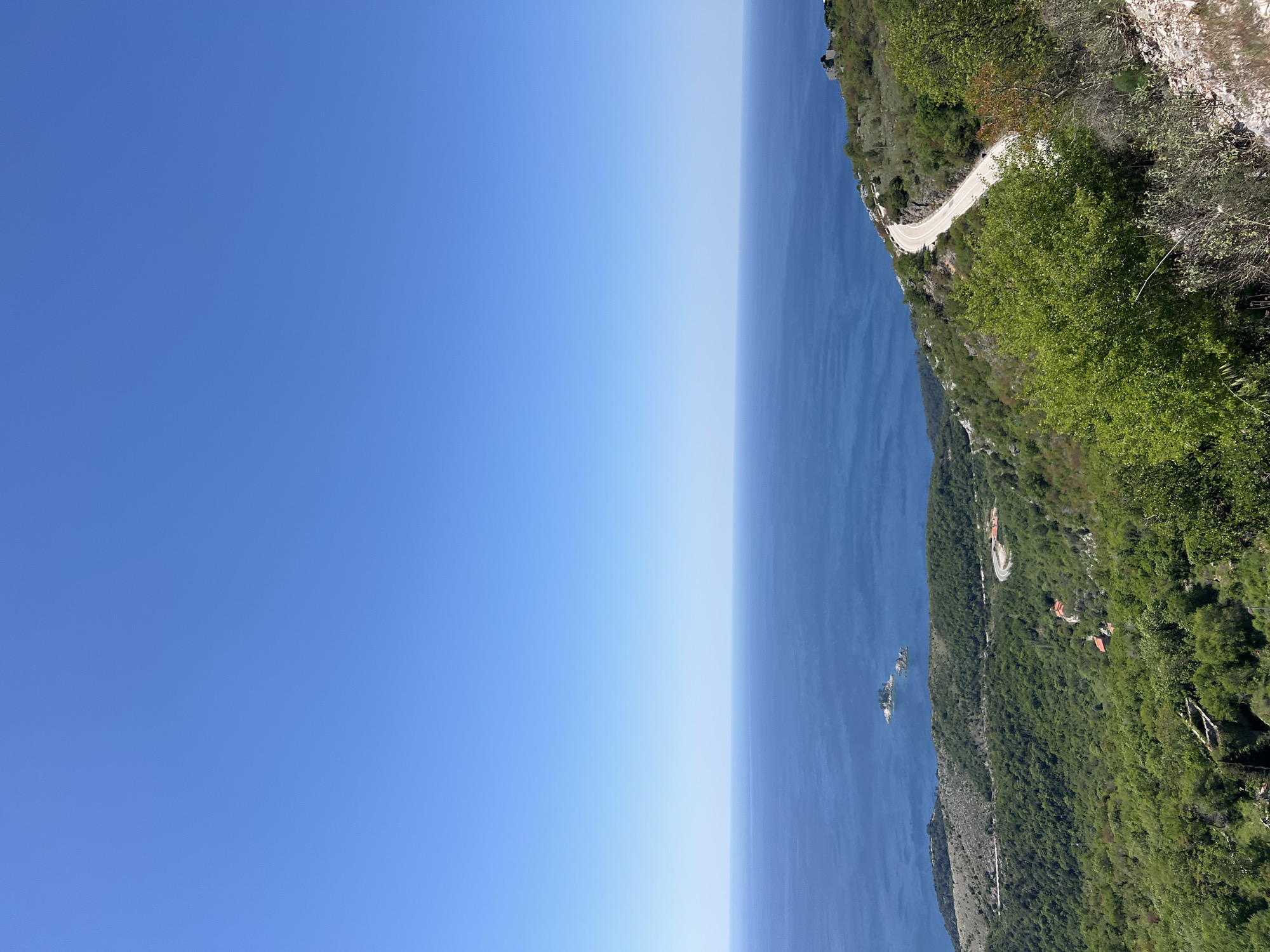 The image shows a coastal view in Novoselje, Montenegro. On the right side, a steep, green hillside with dense vegetation slopes down to the blue water. A winding road is visible along the cliff edge. The left side of the image is a clear, blue sky.