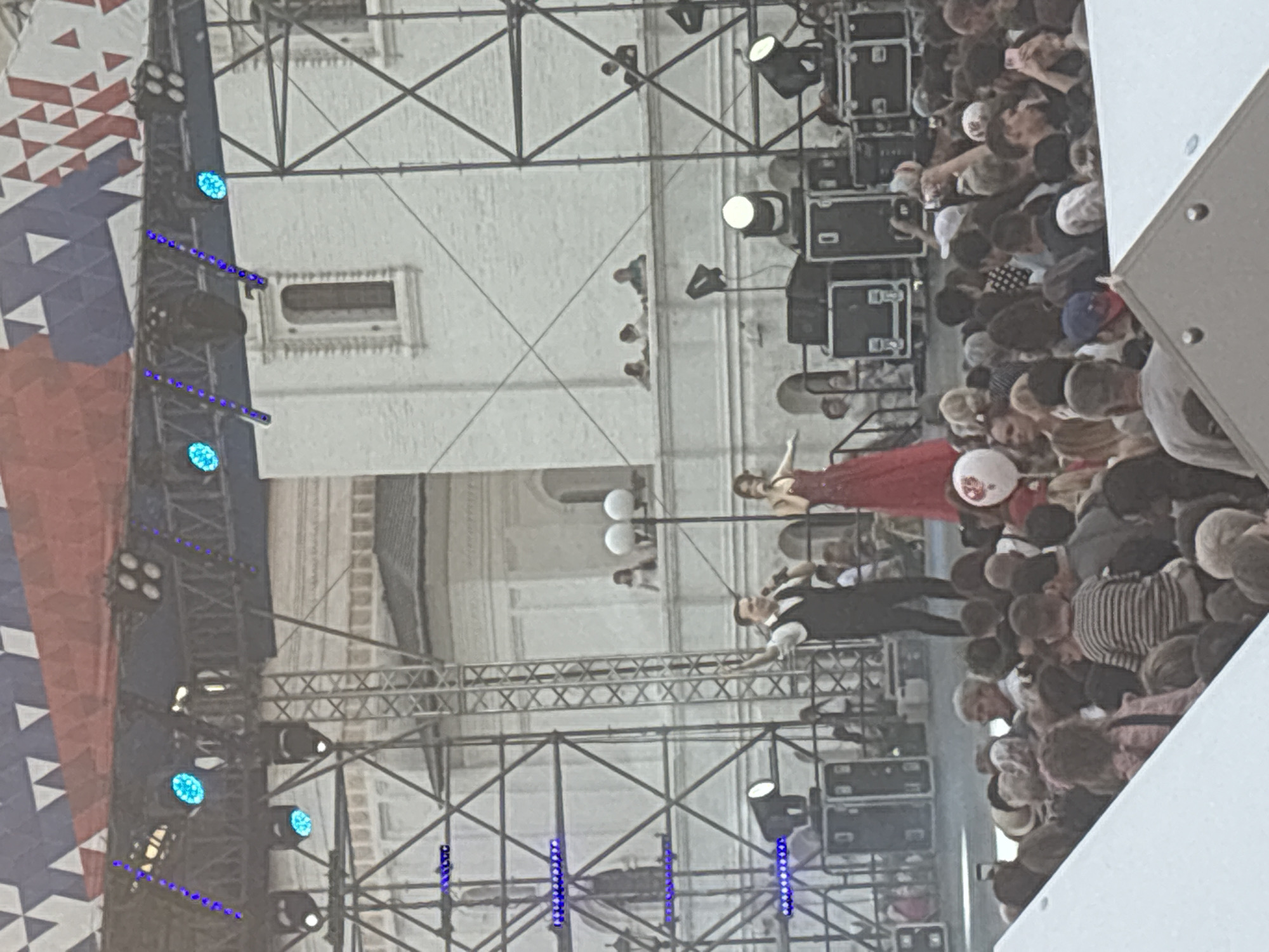 An outdoor concert stage is set up in a city square, with a white building in the background. Two singers, a man in a vest and a woman in a red dress, are performing on stage. They are holding microphones and appear to be singing. The stage is equipped with various lighting and sound equipment. A large crowd of people are gathered in front of the stage, watching the performance. Some people are holding up phones to take pictures or videos. The sky is overcast, and the overall lighting is somewhat dim. The architectural style of the building in the background, with its arched windows and classical detailing, suggests it could be a historical building. The presence of a large crowd and a temporary stage indicates a public event or festival. The overall atmosphere appears to be festive and engaging.