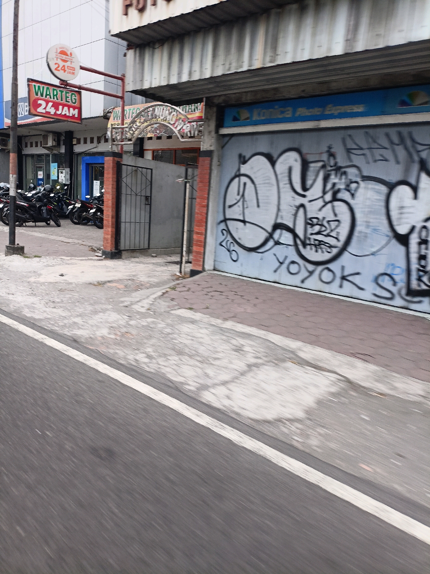 The image depicts a street scene in Magelang, Indonesia, likely in the early morning or late afternoon given the lighting conditions. In the foreground, a grey asphalt road is visible with a white line marking a lane. To the left, a sidewalk area is present with parked motorcycles. A sign with the text "WARTEG 24 JAM" is prominently displayed, indicating a 24-hour food stall. Behind the sign, a building features a storefront with a blue awning and graffiti on its metal shutter. The graffiti includes text such as "YOYOK S" and other stylized lettering. Further to the right, a brick pillar supports part of an overhang. The environment suggests a commercial or roadside area within the city. No people are clearly visible in the image, but the presence of parked motorcycles suggests recent activity.