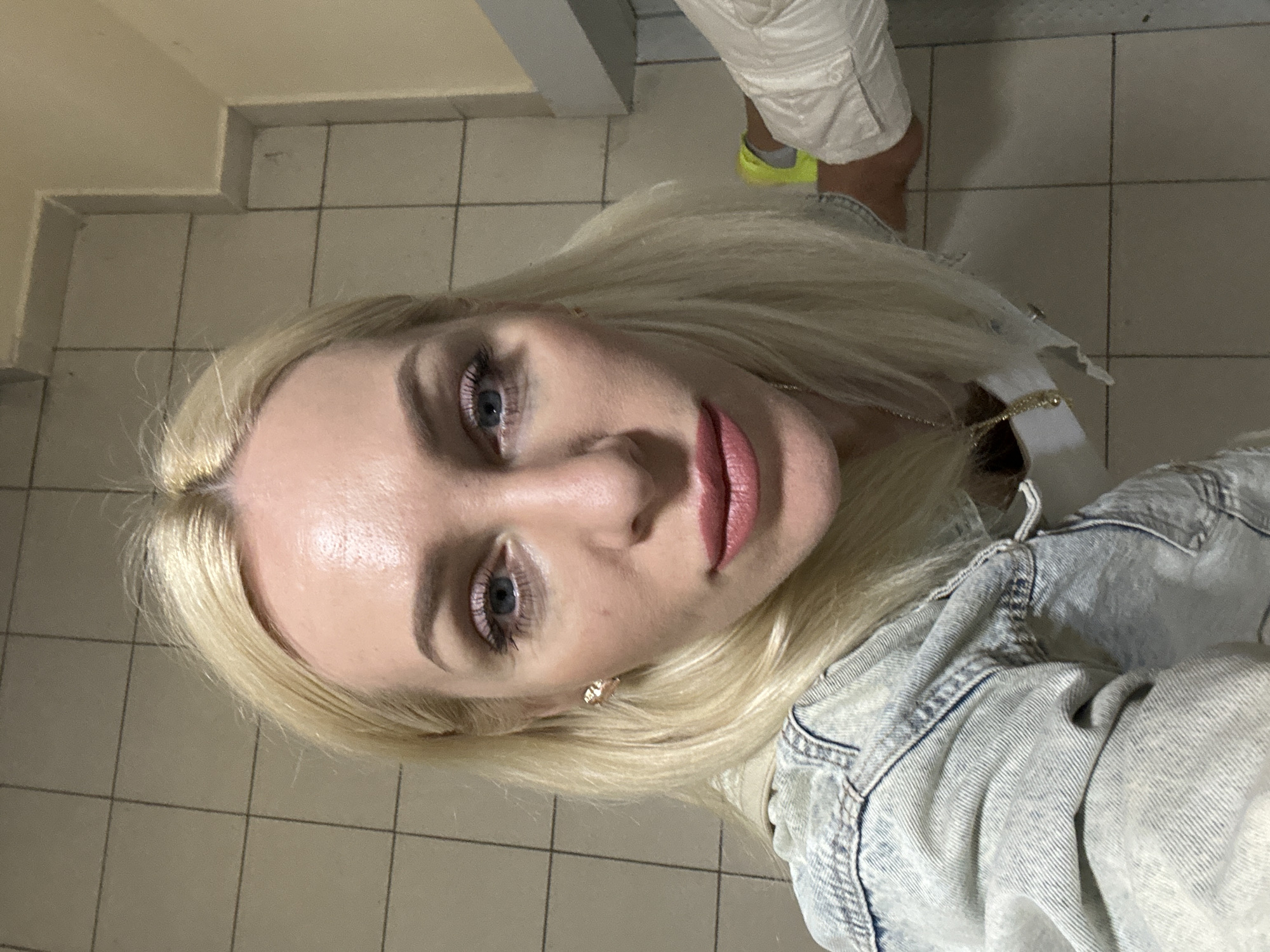 A blonde woman with blue eyes and pink lipstick is posing for a selfie indoors. She is wearing a denim jacket over a white shirt and a gold necklace. The background is tiled with beige square tiles. To the right, a person wearing a white shirt and yellow sneakers is partially visible.