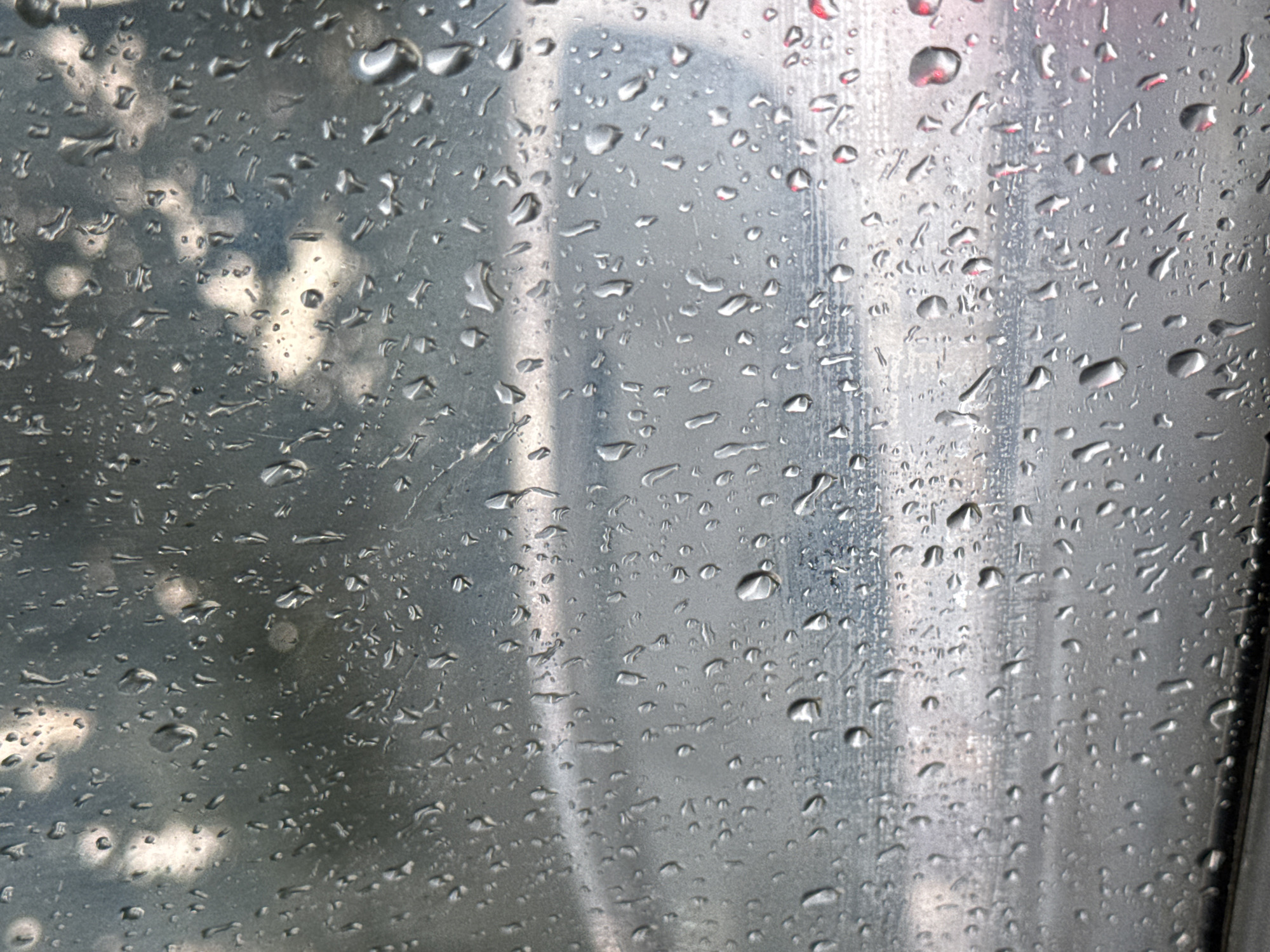 The image depicts a close-up view of a surface covered in raindrops. The raindrops are of various sizes and shapes, some appearing as small, distinct beads, while others are elongated or have merged into larger formations. The surface itself appears to be metallic or glass, with a textured, somewhat reflective quality. Behind the raindrops, a blurred background is visible, suggesting an outdoor environment, possibly a street scene or landscape. The colors in the background are muted, with shades of gray, white, and hints of blue and red, possibly indicating reflections or distant objects. There is no discernible information within the image that can be used to identify the specific location as Tiraspol, Moldova. The scene is static and does not depict any people, actions, or interactions.