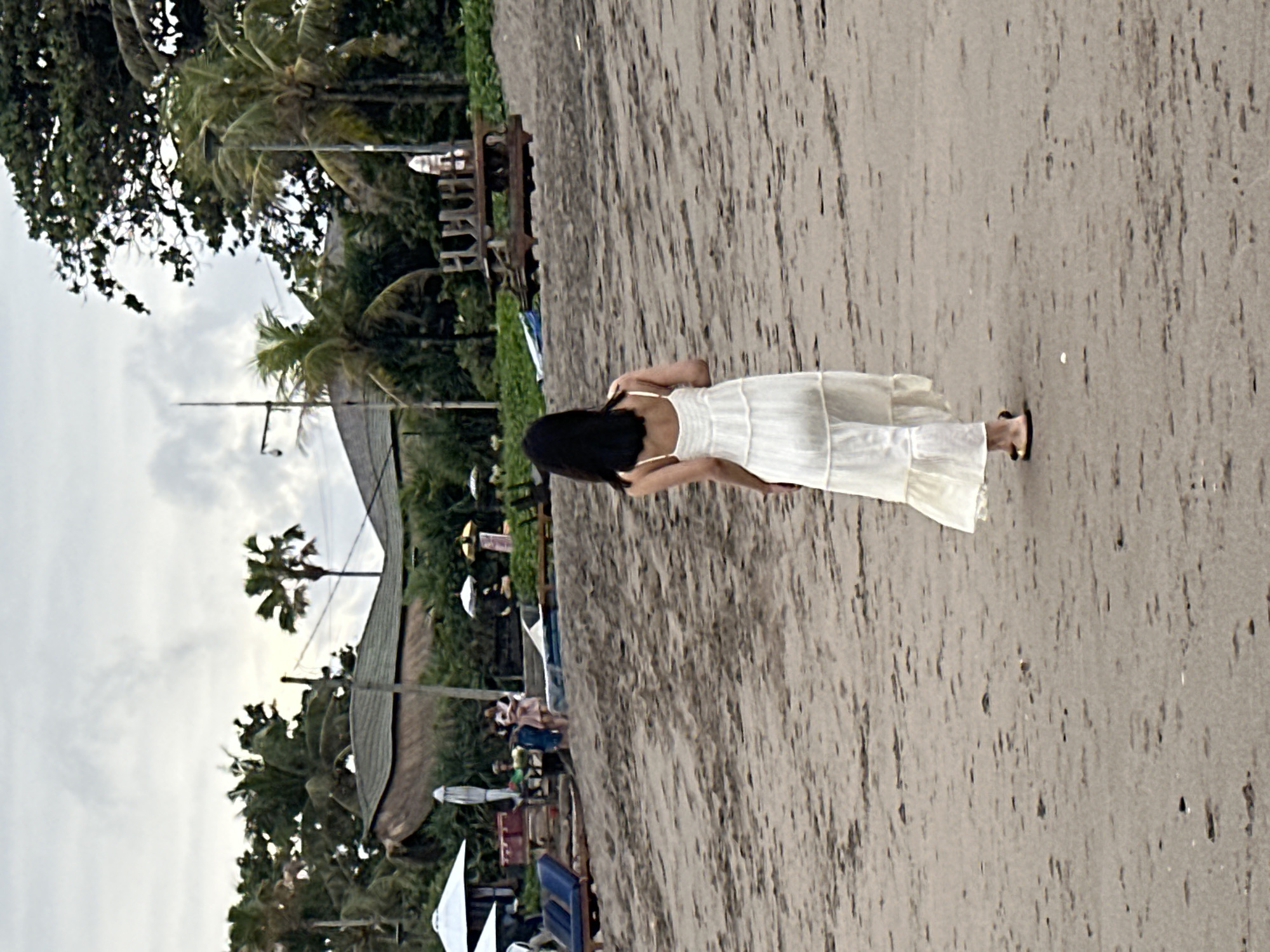 A woman with long, dark hair, wearing a white sundress and sandals, walks away from the camera on a sandy beach. The sky is overcast. In the background, there are palm trees, thatched-roof buildings, lounge chairs, and umbrellas. The scene appears to be a resort or a beachside establishment in North Kuta, Indonesia.