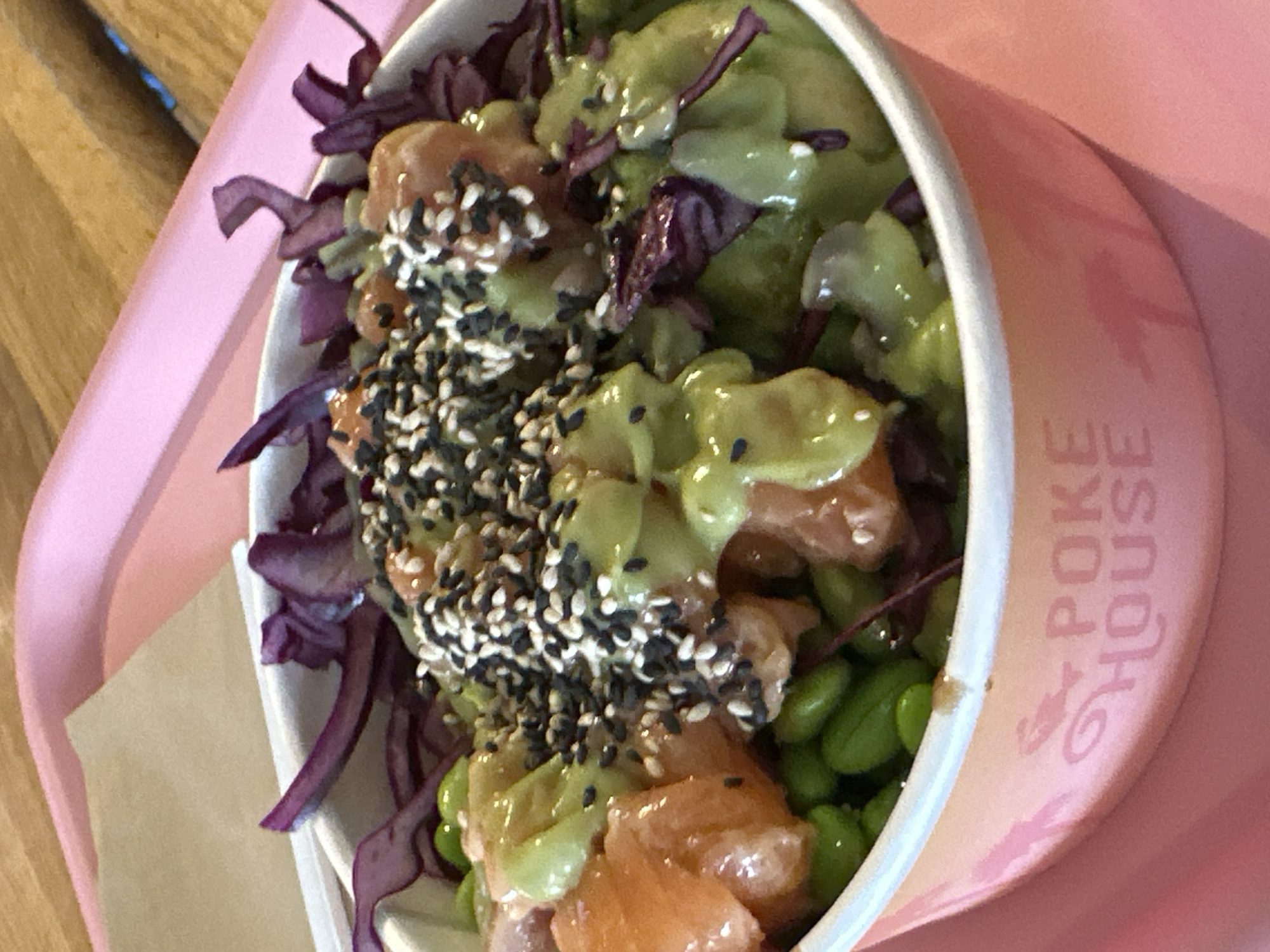 A close-up, eye-level shot shows a poke bowl filled with cubed salmon, avocado sauce, edamame, and purple cabbage, topped with sesame seeds. The bowl is a pale orange with pink lettering that reads "POKE HOUSE". The bowl is sitting on a pink tray. The background is a wooden table. This image was taken at Poke House in Milan, Italy.