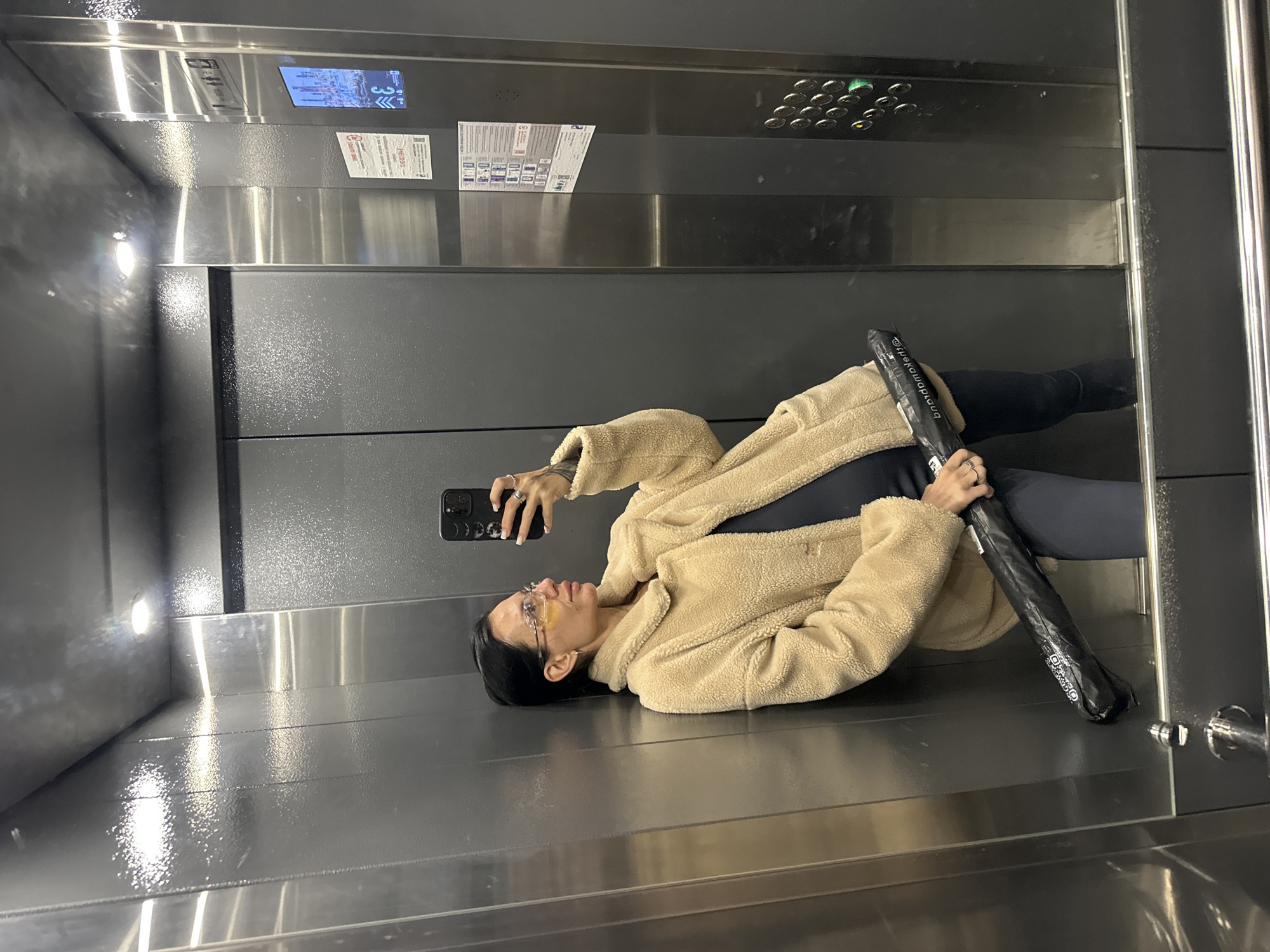 A woman stands in an elevator, leaning against the metallic wall. She is wearing a beige teddy bear coat over dark clothing, and has on glasses. She holds a black umbrella in her right hand and takes a selfie with her phone in her left. The elevator is equipped with buttons, an illuminated panel, and informational signs. The floor and walls are made of polished metal.