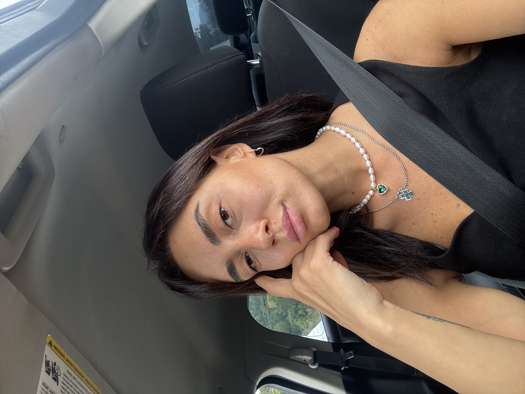 A woman is sitting in the driver's seat of a car. She is looking at the camera, resting her chin on her hand. She is wearing a black tank top, a pearl necklace with a green heart pendant, and a silver chain necklace with a blue cross pendant. She has dark hair and is wearing small hoop earrings. The car interior is visible, including the seatbelt and a warning sticker on the sun visor. Outside the car, trees and a hillside are visible, suggesting a semi-rural or suburban environment. The lighting indicates it is daytime. The location is identified as Tbilisi, Georgia.