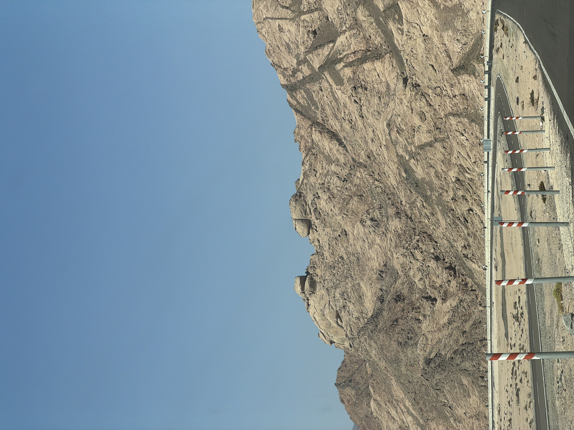 An image captured on a clear day in an unknown location in Egypt showcases a desert landscape dominated by arid, rocky mountains under a vast blue sky. A winding asphalt road cuts through the scene, bordered by red and white striped poles and a concrete barrier with directional signs. The mountains, a mix of tan and grey rock formations, show signs of erosion and are largely devoid of vegetation. The road appears to be an infrastructure project designed to traverse this rugged terrain.