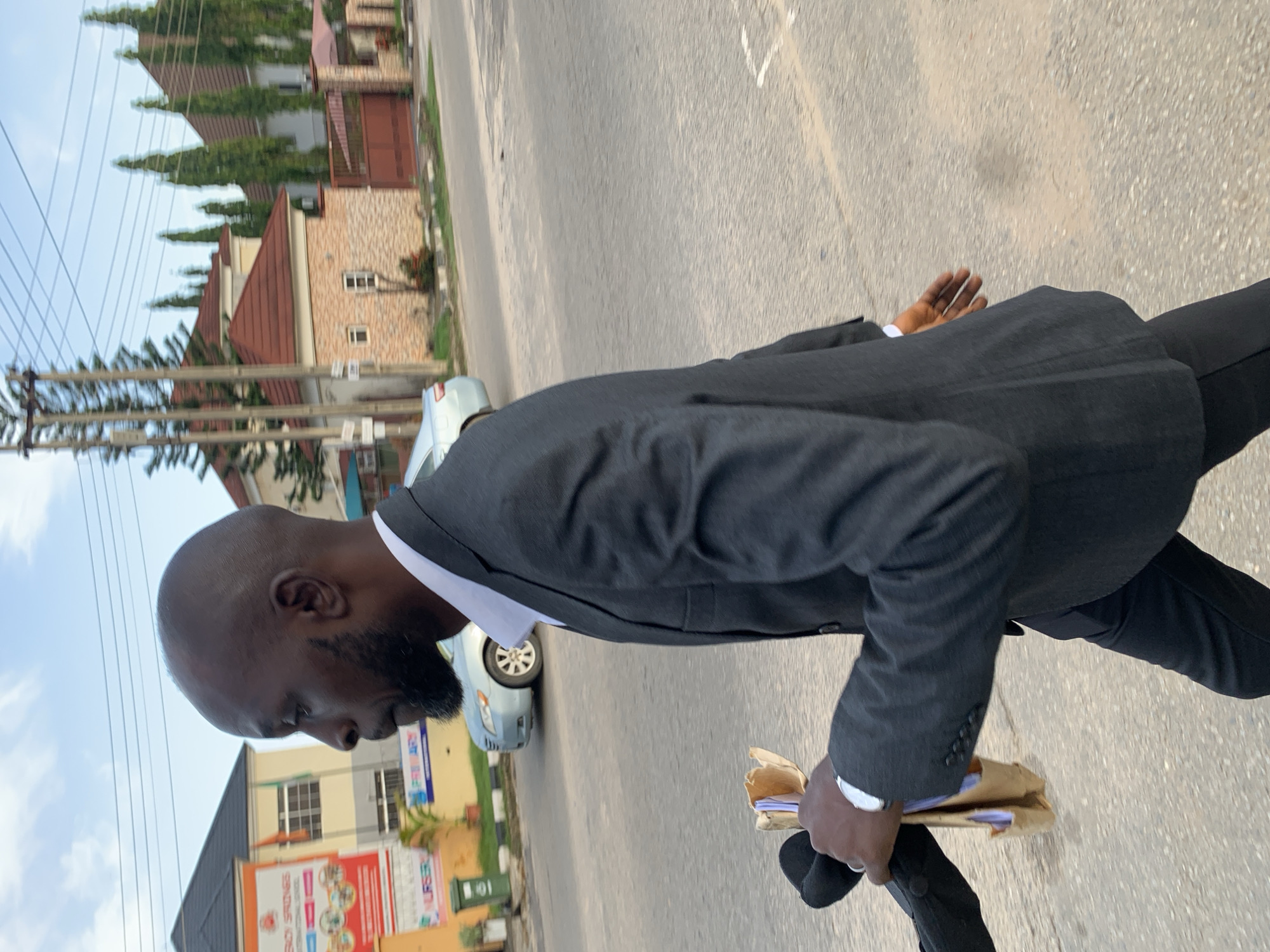 A man wearing a dark suit and white shirt walks on a paved street in Abuja, Nigeria. He has a shaved head and a beard. He is holding a brown folder in his right hand, and a black object, possibly a bag, in his left hand. In the background, there are houses with red roofs, trees, a silver car, and a banner for "Springs International School". The sky is cloudy.