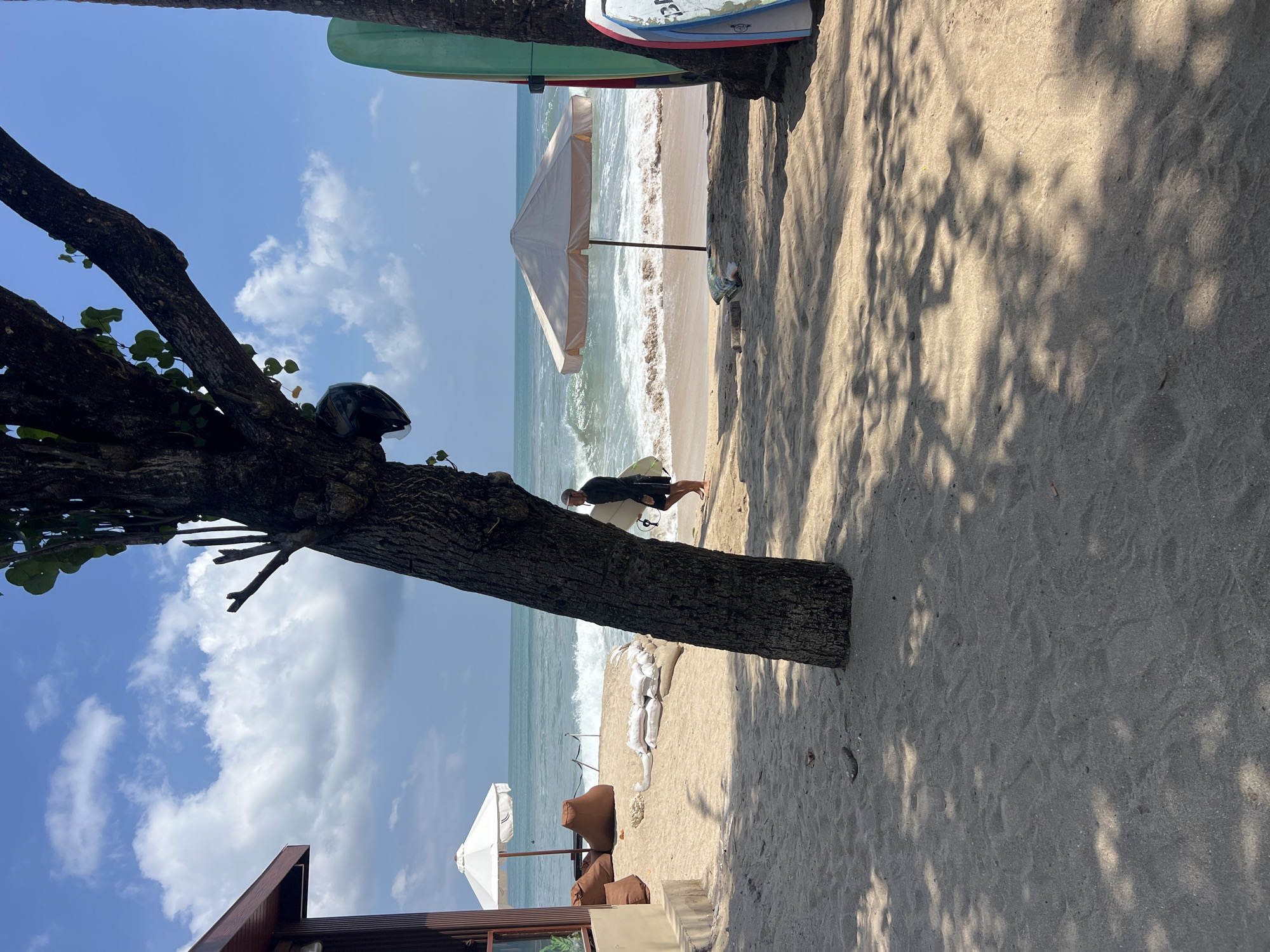 A beach scene in Kuta, Indonesia, under a bright, sunny sky with scattered clouds. A large tree with a helmet hanging from a branch dominates the foreground, its shadow cast across the sand. To the left, a part of a building with a brown, wooden exterior is visible, alongside a white beach umbrella and several brown beanbag chairs. In the middle ground, a person wearing dark clothing walks along the wet sand near the ocean, carrying a white surfboard. Waves are breaking on the shore. Further to the right, two surfboards, one green and one with blue and red accents, are leaning against a palm tree. A light-colored beach umbrella is also set up on the sand near the water. The sand is light brown and appears undisturbed in most areas, with some footprints visible.