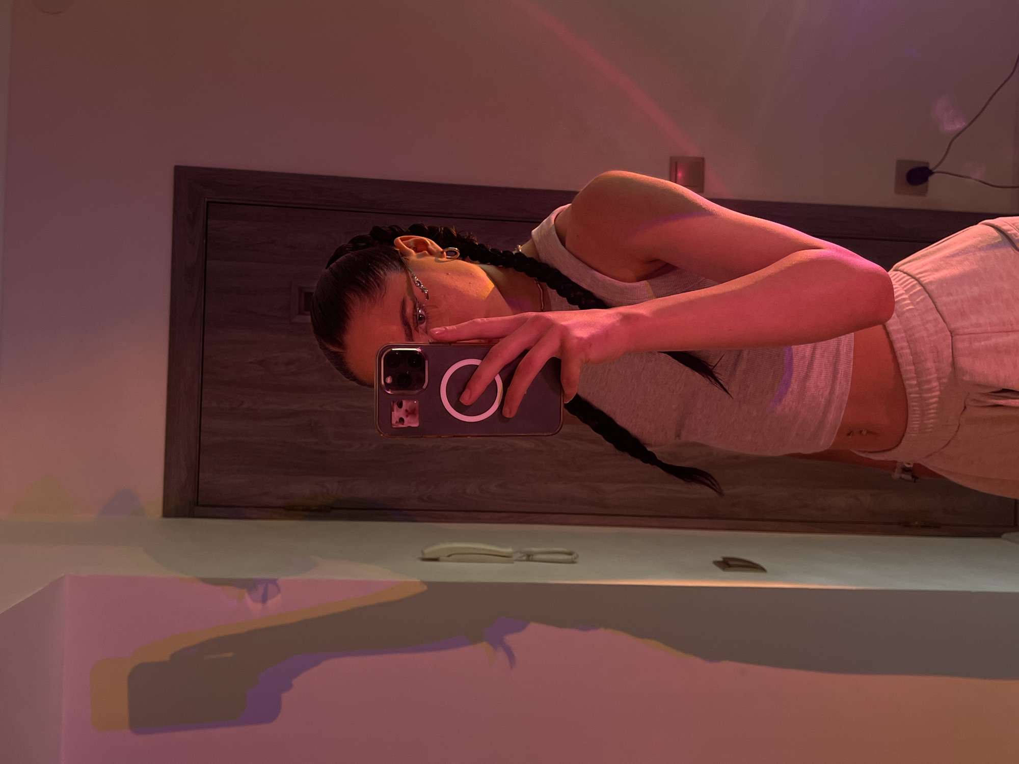 The image is a full-body mirror selfie taken by a young woman indoors. She is wearing a light gray tank top and light gray sweatpants. Her dark hair is styled in two braids that fall over her shoulders. She is wearing distinctive rhinestone-embellished glasses. In her right hand, she holds a smartphone, which is capturing the reflection. The phone case has a small image of a cat's face and a circular magnetic charging accessory. The background shows a closed door with a dark wooden frame and a wall illuminated by colored lights, casting shadows on the wall to the left. The lighting suggests an artificial light source, possibly a colored lamp or LED strip, creating a pink and orange hue in the environment. The overall scene appears to be a bedroom or a similarly private indoor space.
The image does not contain any information that can be used to determine the location in the city of Ternopil, Ukraine.