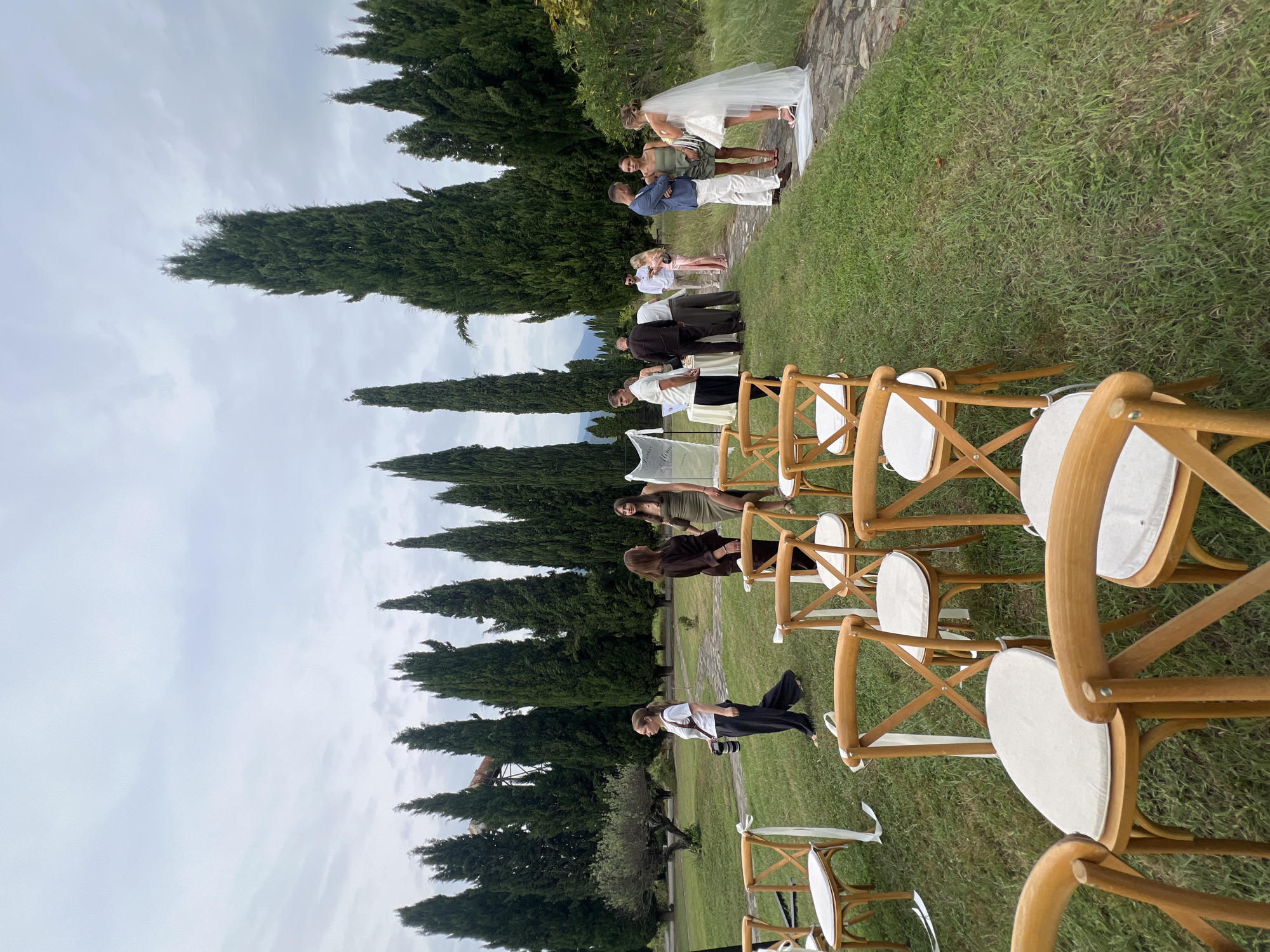 The photograph captures an outdoor wedding ceremony set in Saniore, Georgia, with a backdrop of tall, slender cypress trees. In the foreground, rows of empty wooden chairs with white cushions are arranged on a lush green lawn, each adorned with a white ribbon. A stone pathway runs along the right side of the image.
Several groups of people are visible scattered across the lawn and near the pathway. On the left, a woman in black pants and a white t-shirt is walking away from the camera, carrying a camera bag. Near the center, a group of guests are standing and mingling. On the right, a bride in a white wedding dress and veil stands with a groom, who is wearing a blue suit. Other guests are interacting in small clusters, some near a table draped with a white cloth. The sky is overcast, suggesting a temperate or humid climate. The overall scene depicts a serene and elegant outdoor wedding venue.