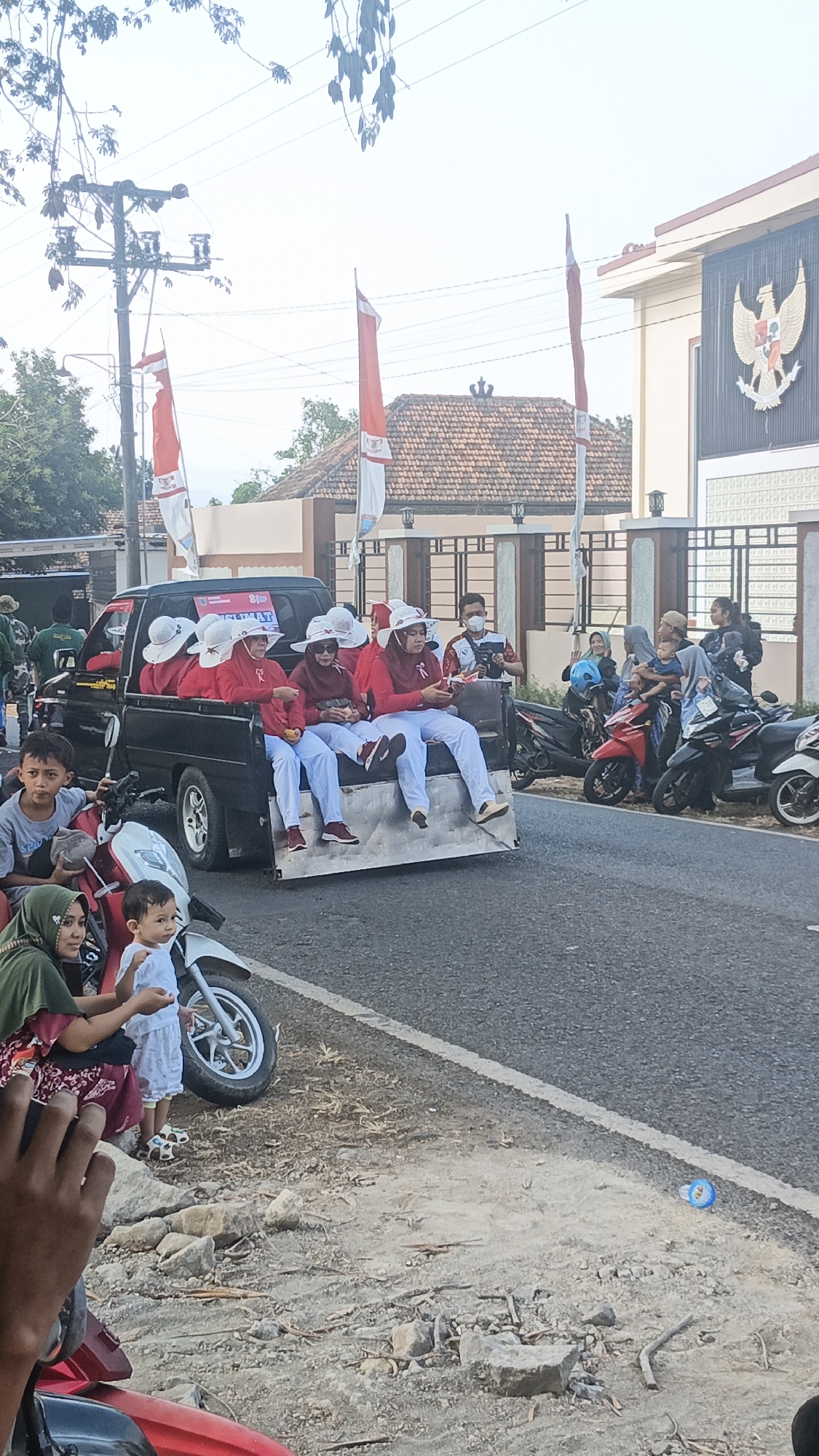 In the city of Kandangmas, Indonesia, a group of people are participating in a parade. The parade features a black pickup truck carrying several individuals dressed in red shirts and white hats. The truck is adorned with Indonesian flags. In the foreground, a young boy sits on a motorcycle, looking towards the truck. Beside him, a woman in a green headscarf holds a small child. The scene takes place on a road, with buildings and trees visible in the background. Several other motorcycles are parked along the roadside, and some spectators are observing the parade.
