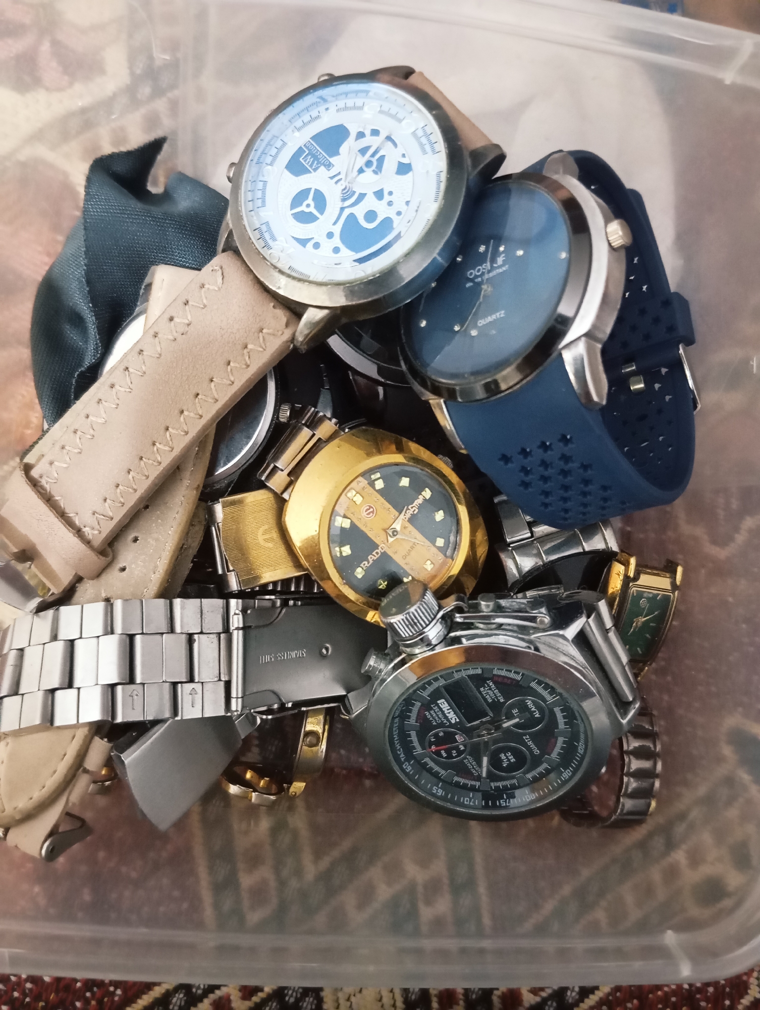 An overhead, eye-level, medium shot shows a pile of wristwatches in a clear plastic container. The watches are of various styles, colors, and materials. In the foreground, a silver metal watch with a black face is prominent, with intricate markings and sub-dials. To its left, a tan leather-banded watch is partially visible, with visible stitching. Above these, a gold-tone watch with a dark face and gold accents is nestled amongst other timepieces. Towards the top of the frame, a watch with a blue dial and a blue rubber strap is clearly visible. Another watch with a white skeletal dial and blue accents is situated at the top left. Several other watches and watch parts are partially obscured within the container. The background is blurry, showing the textured surface of the container and a patterned fabric beneath it.
