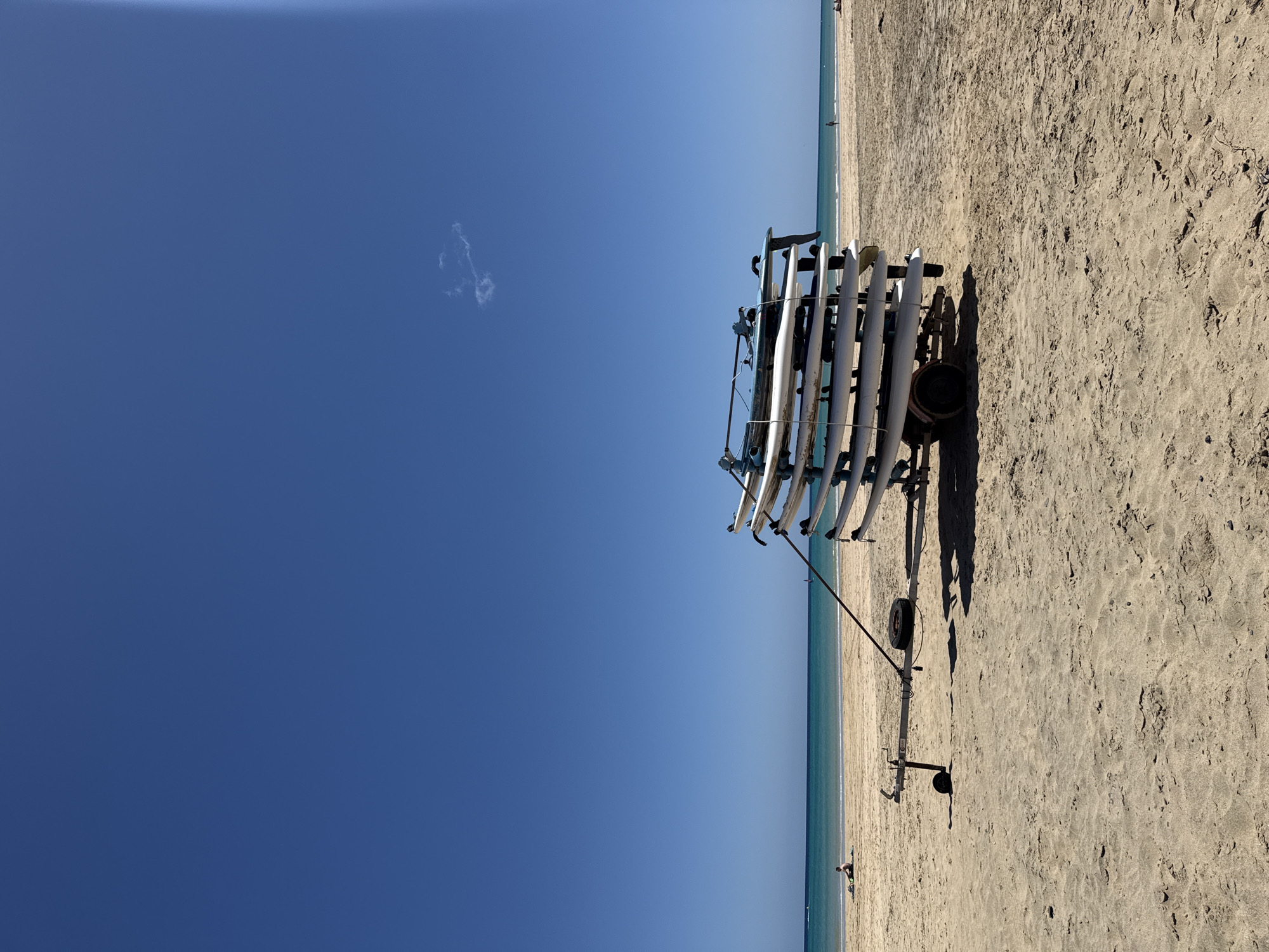 The image captures a sunny scene on a beach in Pájara, Spain. A tall, wheeled cart is loaded with numerous white surfboards or paddleboards, stacked horizontally. The cart casts a shadow on the light-colored sand. Beyond the cart, a calm, light blue sea stretches to the horizon, where it meets a lighter blue sky. The sky is mostly clear, save for a small, puffy white cloud. In the distance, people can be seen on the beach and in the water.