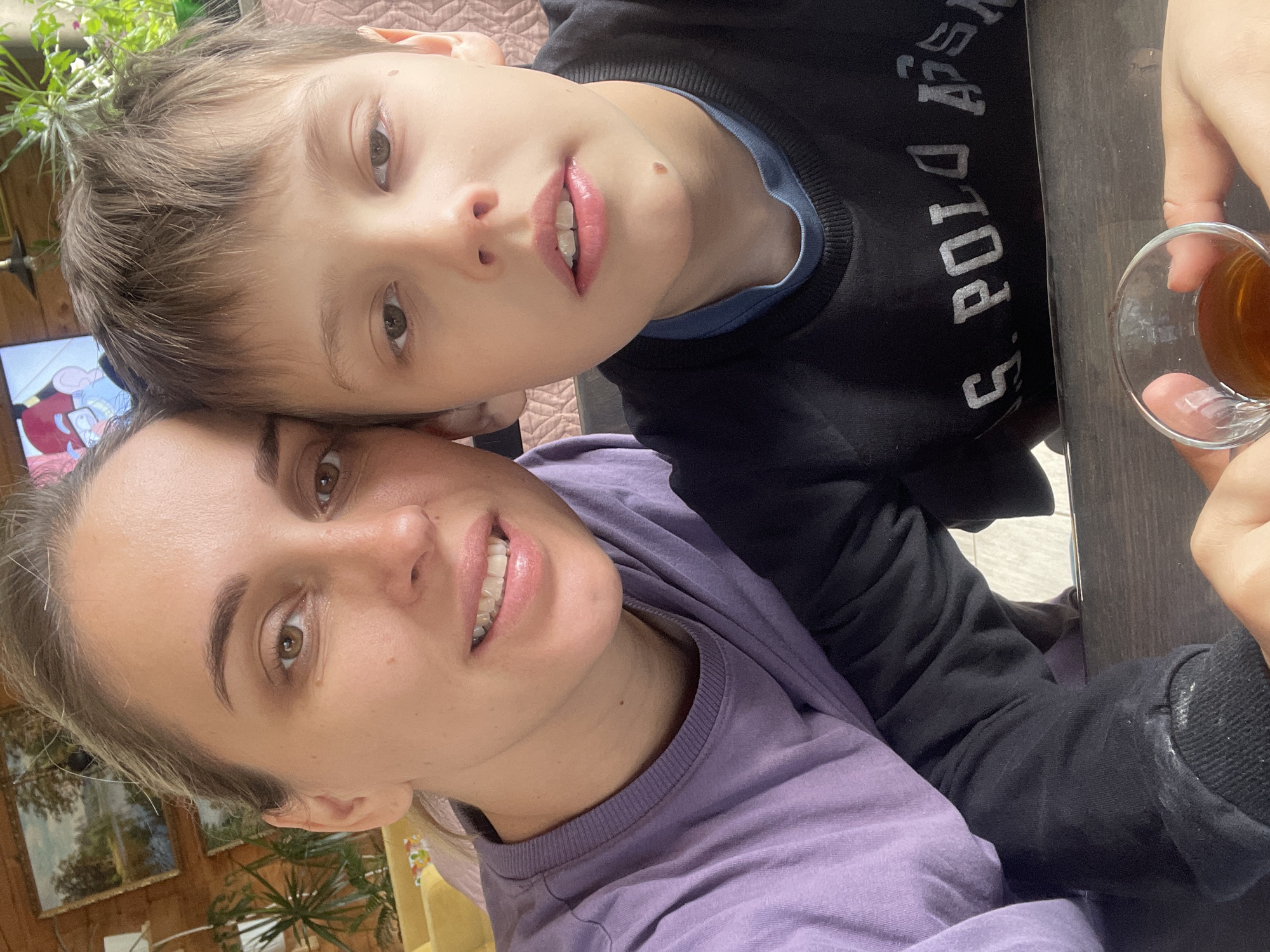 The image shows a selfie of a woman and a child. The woman is wearing a purple shirt and has light brown hair. The child, who appears to be a boy, is wearing a black sweatshirt with "S. POLO ASSN." printed on it. They are close together, likely indoors. In the background, there's greenery, possibly plants, and glimpses of wall decor suggesting a home environment. A glass of liquid, possibly tea, is visible in the foreground. The image may have been taken in Chaikovsky, Russia.