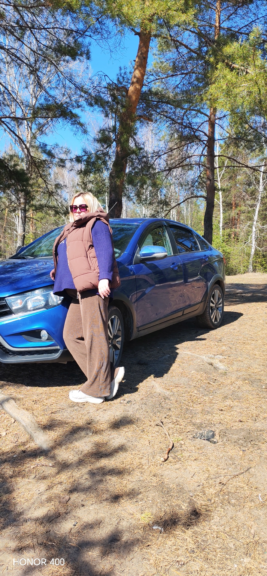 A woman stands in a forest setting next to a blue Lada Vesta car. The woman has blond hair, wears sunglasses, a brown vest, a blue sweater, brown pants, and white sneakers. The ground is covered in dry pine needles. Trees surround the car. The text "HONOR 400" is in the lower-left corner of the image. The location is likely near Noviy, Russia, based on the car model, but this cannot be confirmed from the image alone.