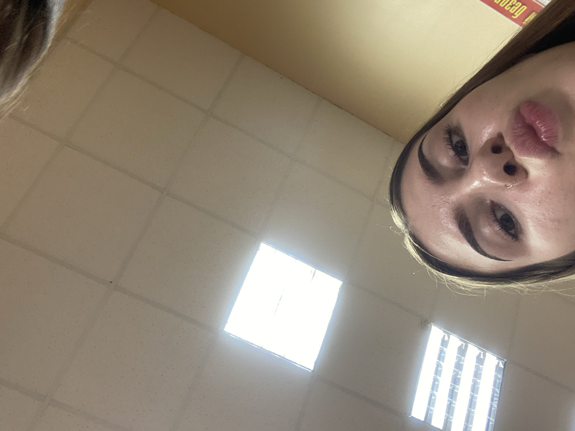The image is an upward-facing selfie taken by a young woman. She has brown hair and is wearing a nose ring. Her lips are pursed, and her eyes are looking upwards. She appears to be indoors, with a drop ceiling visible in the background. There are fluorescent lights in the ceiling. A yellow and red sign with Cyrillic lettering is partially visible on the right side of the frame. The location is identified as Дьяково, Russia.