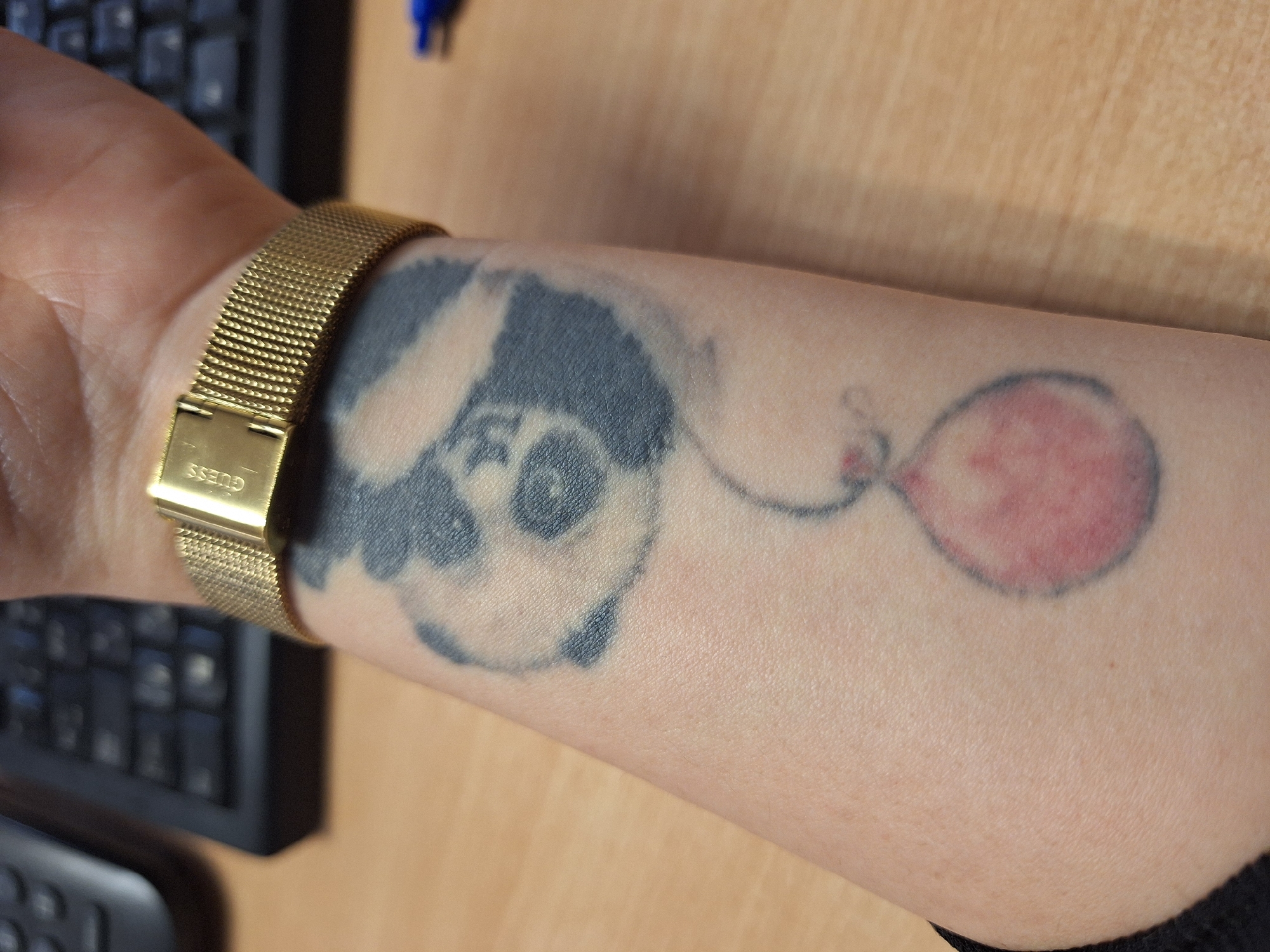 An arm with a faded tattoo of a panda holding a balloon is resting on a desk next to a keyboard. The arm has a gold-colored watch with the brand name "GUESS" visible on the clasp. The desk has a light brown surface. The location is not discernible from the image; there is no information suggesting that the image was taken in Varna, Bulgaria.