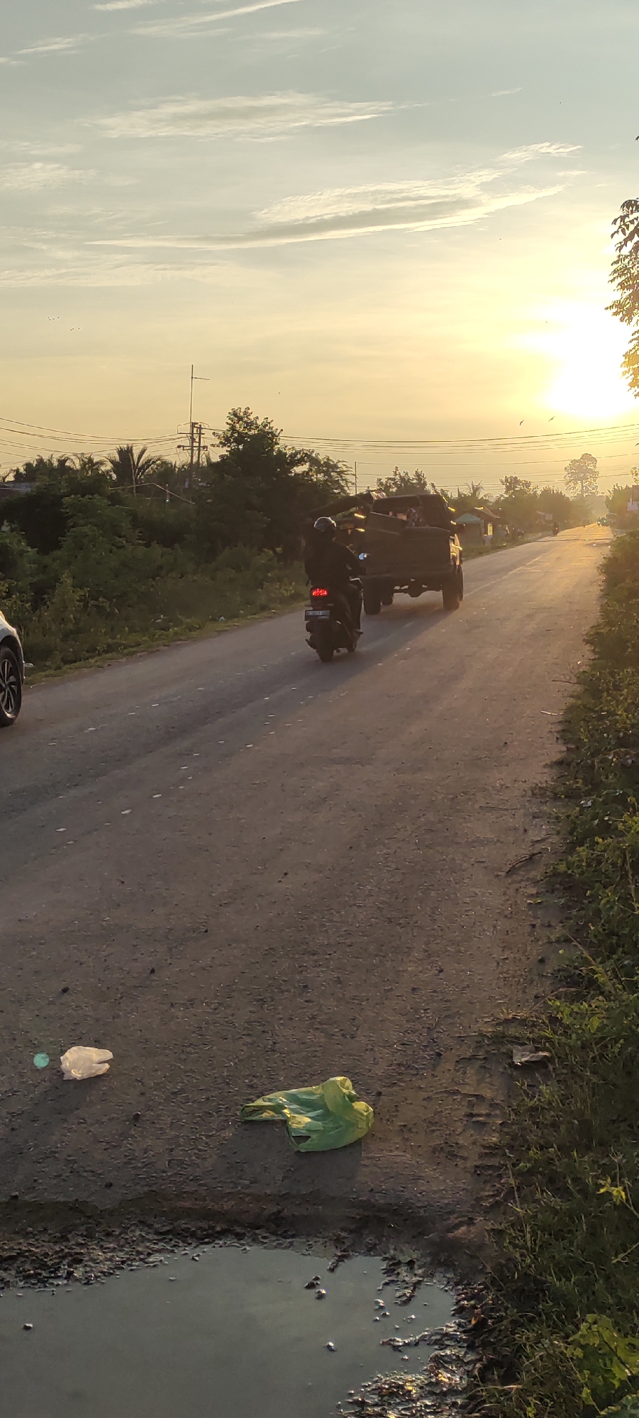 An outdoor, vertical, medium shot captures a rural road in Blangmajron, Indonesia during sunset. The sun is setting to the right of the frame, casting a golden glow across the sky and illuminating the scene with warm light. In the foreground, the asphalt road is marred by a puddle of murky water and scattered litter, including two plastic bags, one white and one green. A motorcycle, with a rider wearing a helmet and dark clothing, travels away from the viewer on the road. Behind the motorcycle, a truck, also carrying what appears to be cargo, moves in the same direction. The roadside is bordered by dense green vegetation and trees, with hints of scattered houses or buildings in the distance. Overhead power lines are visible against the pale, cloud-streaked sky. The overall impression is of a quiet, everyday scene on a rural road in Indonesia at dusk.