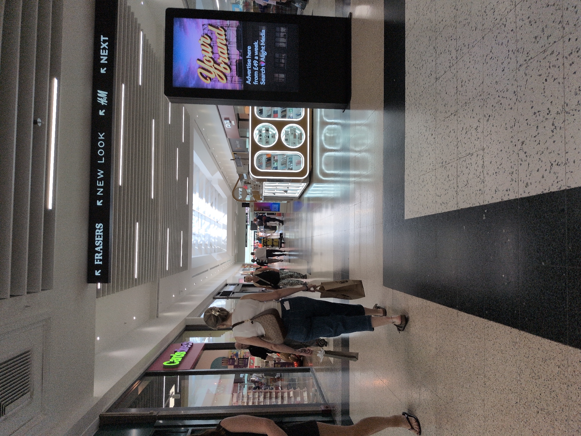 The image shows the interior of a shopping mall in Telford, United Kingdom. In the foreground, a woman with blonde hair pulled back is walking away from the camera, carrying shopping bags. She is wearing a white tank top and blue jeans. To her left, another woman is partially visible, wearing a black dress. In the background, the mall is illuminated by overhead lighting and the storefronts of various shops are visible, including signs for "FRASERS", "NEW LOOK", "H&M", and "NEXT". A digital advertising screen displays the text "Your Brand" and "Advertise here from £49 a week. Search Alight Media". Several other shoppers are visible in the distance. The mall floor is tiled with a speckled pattern and a black stripe.