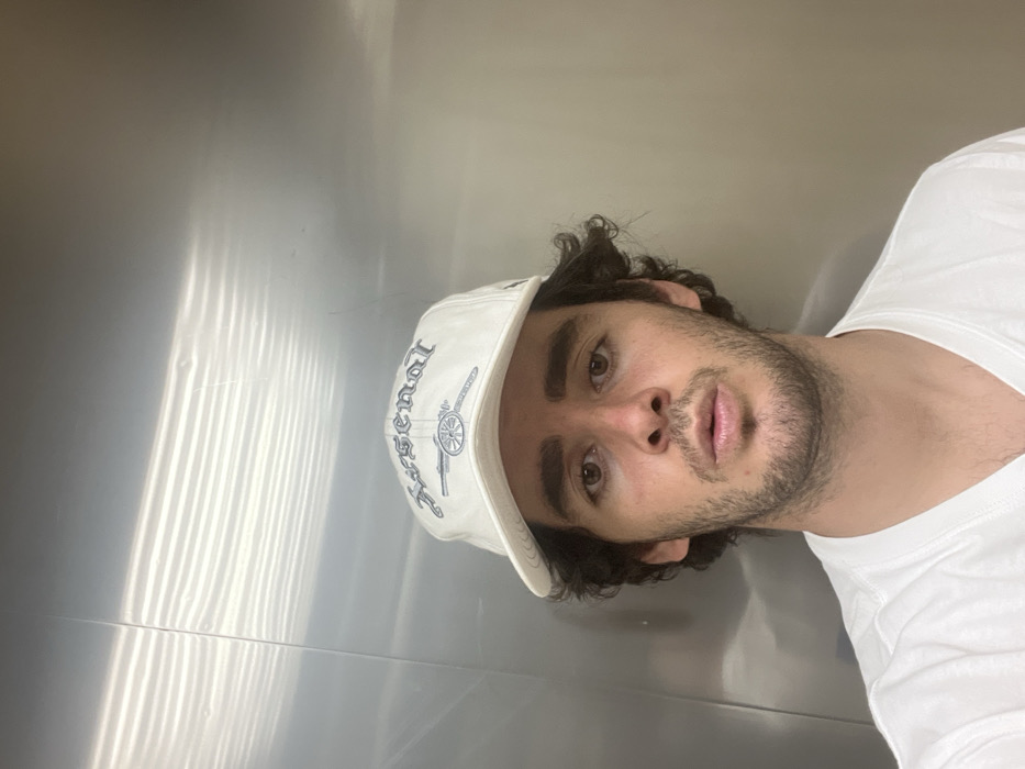 A young adult male is depicted in a close-up, frontal view within an enclosure featuring brushed metallic walls, consistent with an elevator interior, in Unknown, Unknown. The subject has dark, curly hair, brown eyes, and facial stubble, maintaining a direct gaze with his lips slightly parted. He wears a white V-neck t-shirt and a white baseball cap. The cap is embroidered with light blue/gray "Arsenal" text in a gothic script above a cannon motif. The metallic background exhibits horizontal striations and reflections from an overhead light source.