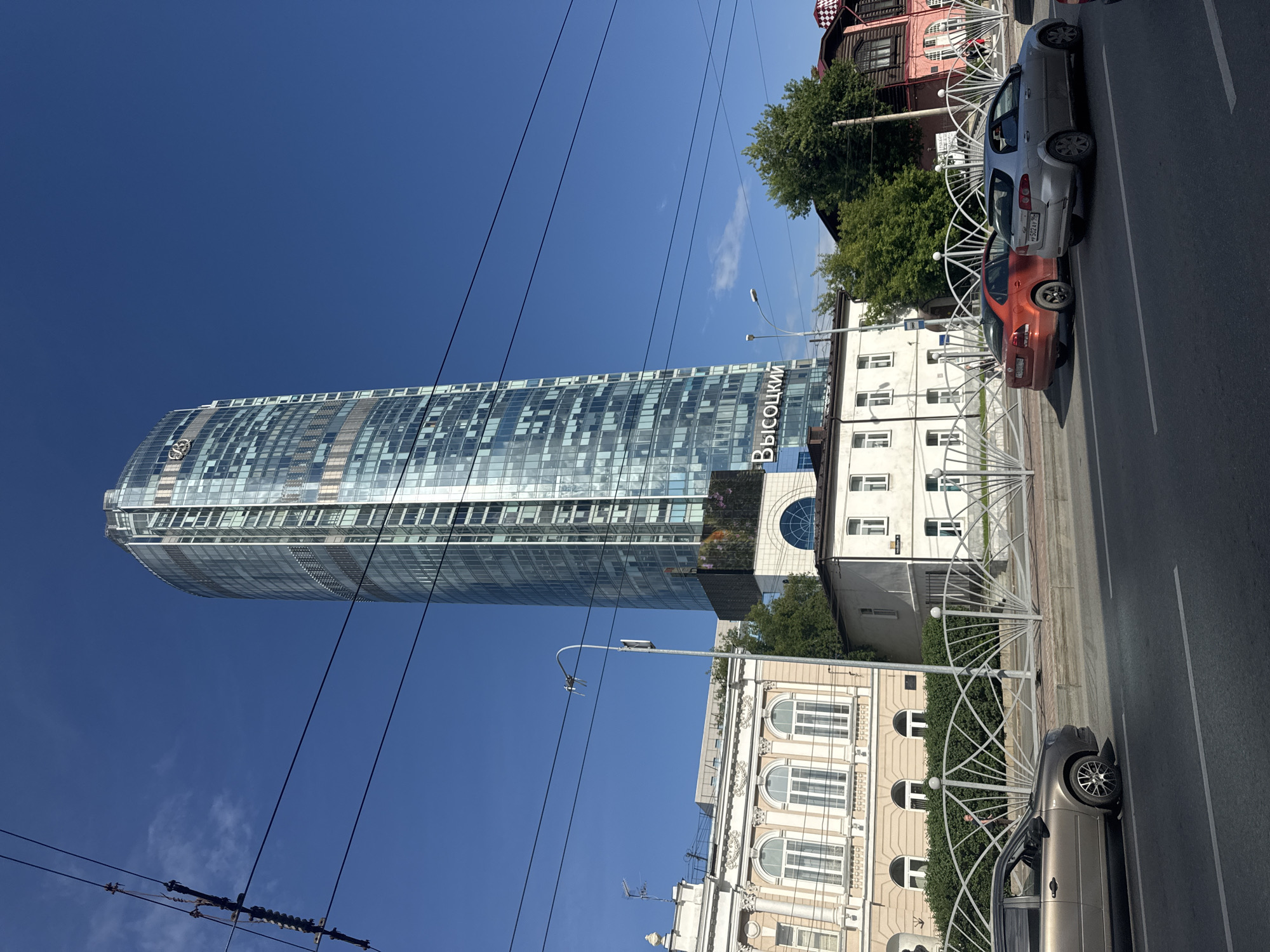 This image, taken in Yekaterinburg, Russia, features the "Vysotsky" skyscraper, a prominent modern building with a glass facade, set against a clear blue sky. The skyscraper is the central subject, with its name, "Высоцкий", clearly visible in white lettering on its side.
The scene depicts an urban environment with a street running alongside the skyscraper. Several cars are parked along the street, including a silver sedan and an orange hatchback. The buildings in the foreground and mid-ground display a mix of architectural styles, with one building featuring classical detailing and arched windows, and another a more contemporary design with a rounded window.
The time of day appears to be daytime, judging by the bright sunlight casting shadows and illuminating the scene. The weather is clear, with only a few wisps of clouds visible in the sky. Electrical wires and a lamppost are also present, indicating urban infrastructure.
The overall impression is a snapshot of city life, showcasing the juxtaposition of modern architecture with older buildings and everyday urban elements. There are no people prominently featured in the image, and no specific activity or event is clearly taking place, suggesting a general view of the street and buildings.