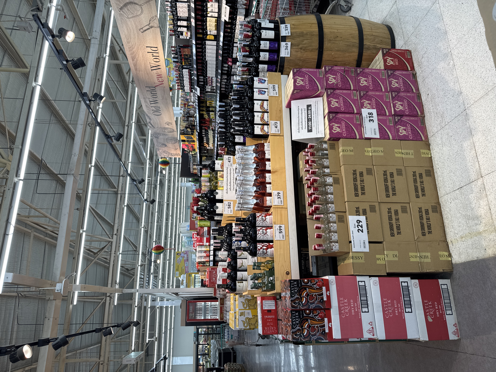 The image depicts the alcohol section of a grocery store, likely in Thong Sala, Thailand. The aisles are lined with shelves stocked with wine, spirits, and pre-mixed alcoholic beverages. Overhead, a banner distinguishes "Old World" and "New World" wines. There are many bottles of Mont Clair and Berri Estates. Displayed prominently at the end of the aisles are stacks of "Spy Classic" mixed drinks and "Castle Creek" dry red wine. Prices are displayed on nearby shelf-talkers. Boxes of Smirnoff vodka are stacked next to the Spy drinks. A wooden barrel rests atop the stacked wine. The scene includes metal shelves, overhead lighting, and a gray concrete floor.