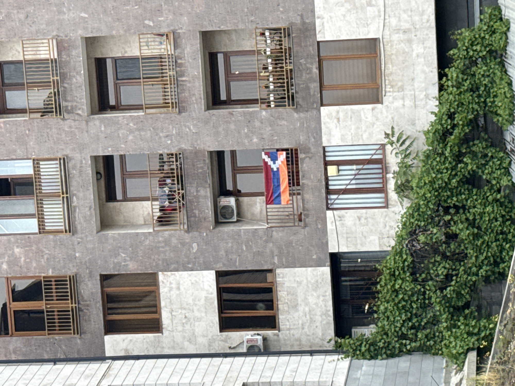 The image shows the facade of a residential building in Yerevan, Armenia. The building has multiple windows, some with metal bars. An Armenian flag is visible hanging from one of the balconies. There are air conditioning units installed on some of the balconies and walls. A large green vine covers a section of the building's exterior.