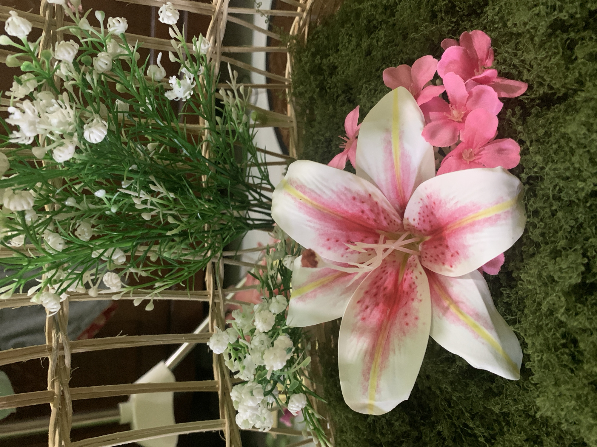 The image shows a close-up view of artificial flowers arranged on green moss. The primary focus is a large, white lily with pink streaks and yellow accents. To the left of the lily are small, white, clustered flowers, resembling baby's breath, and to the right, several smaller pink flowers are visible. The arrangement is placed within what appears to be a woven basket or container. The background consists of a blurred, wooden structure. There is no identifiable location within Kuala Pembuang, Indonesia, or any indication of people or actions. The scene is static and decorative.