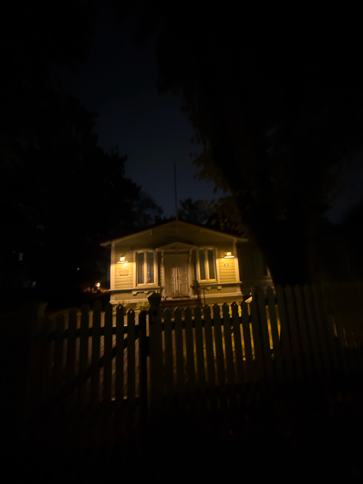 The image shows a light-colored, two-story house at night, viewed from across a white picket fence. The house has several windows illuminated from within, and a porch light shines brightly above a window. The yard is dark and appears to be surrounded by trees, contributing to the nighttime ambiance. The picket fence is partially illuminated by the house lights, casting long shadows. The sky above is a deep, dark blue, typical of nighttime. The scene appears to be a residential area. This photograph was taken in Tallinn, Estonia.